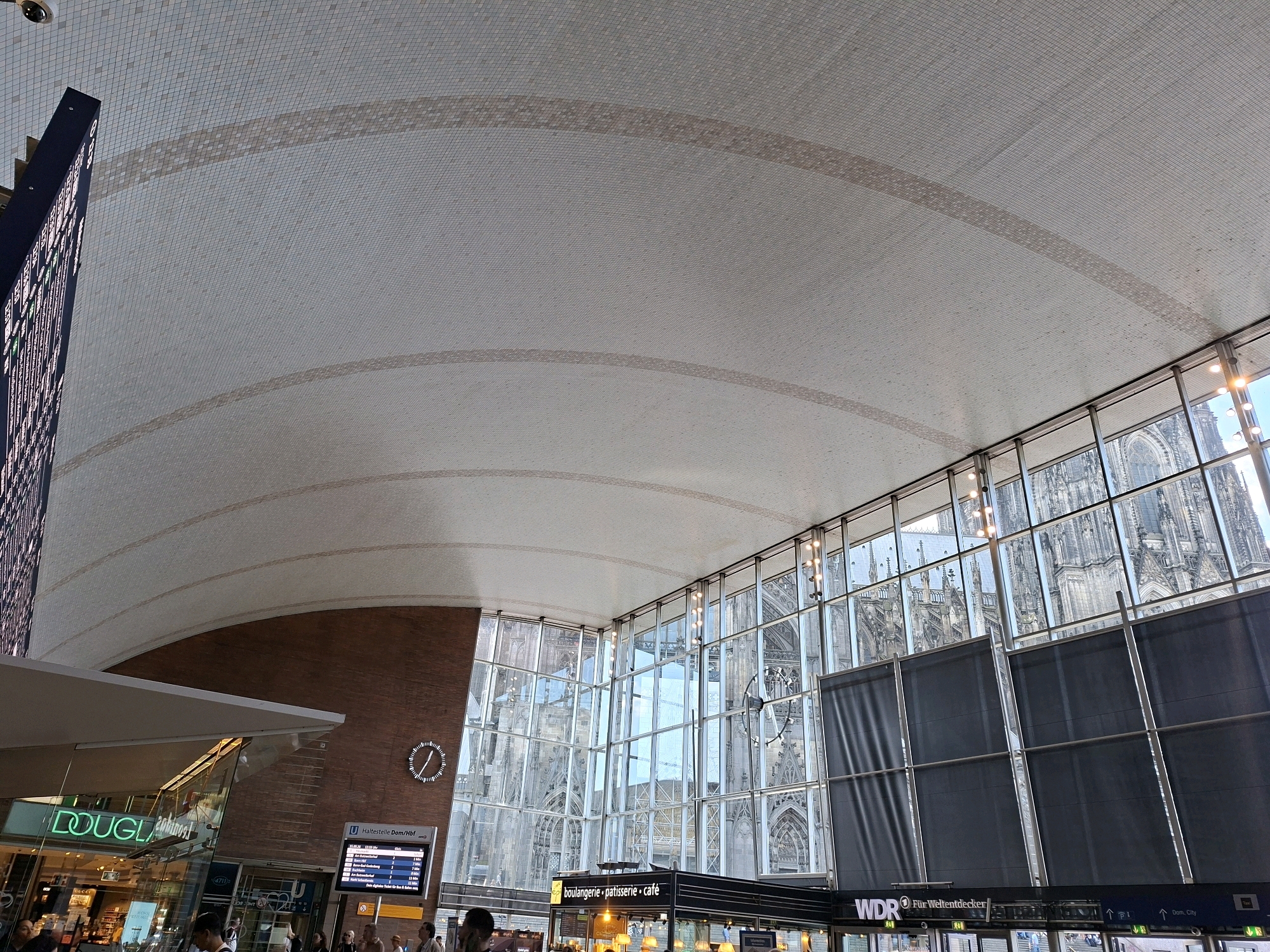 This is an interior photograph taken inside a modern building with a high, vaulted, tiled ceiling. Large glass windows line one wall, offering a view of the Cologne Cathedral in the background, which is a prominent Gothic cathedral located in Cologne, Germany. Through the windows, the cathedral's spires and ornate architecture are visible. Inside the building, there is a large digital departure board displaying train information, a clock on a brick wall, and storefronts, including one labeled "DOUGLAS." Several people are present, some walking, and others appearing to be waiting. The overall scene depicts a bustling interior space within a city, with the historic cathedral serving as a dramatic backdrop.
