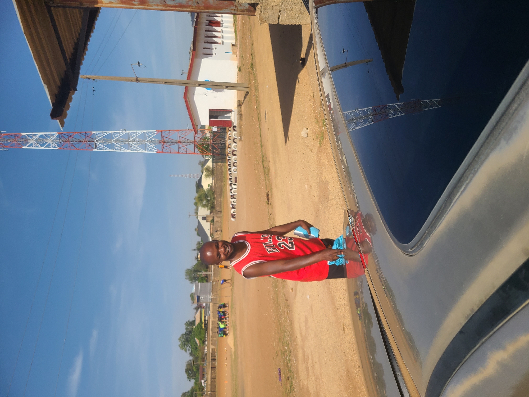 The image depicts an outdoor scene in what appears to be a community area, set against a bright, clear blue sky, likely in Jos, Nigeria. The central subject is a smiling man, dressed in a red basketball jersey featuring "BULLS" and the number "23" on the front. He is actively engaged in cleaning the side of a dark-colored vehicle, possibly a car or SUV, using a light blue cloth. His reflection, along with that of the surrounding environment, is visible on the car's polished surface.
The setting is characterized by an expansive area of reddish-brown dirt ground. In the background, a series of low-rise white buildings with red accents or roofs can be seen. A prominent feature is a tall, red and white lattice-patterned tower, likely a telecommunications or power tower, standing tall against the sky. Overhead, power lines stretch across the frame, supported by wooden utility poles. In the upper left, a section of a corrugated metal roof structure is visible. A line of white-painted tires forms a boundary across the mid-ground. The bright sunlight casts visible shadows, suggesting it is daytime, possibly mid-morning or afternoon. The man's cheerful expression indicates a positive demeanor.
