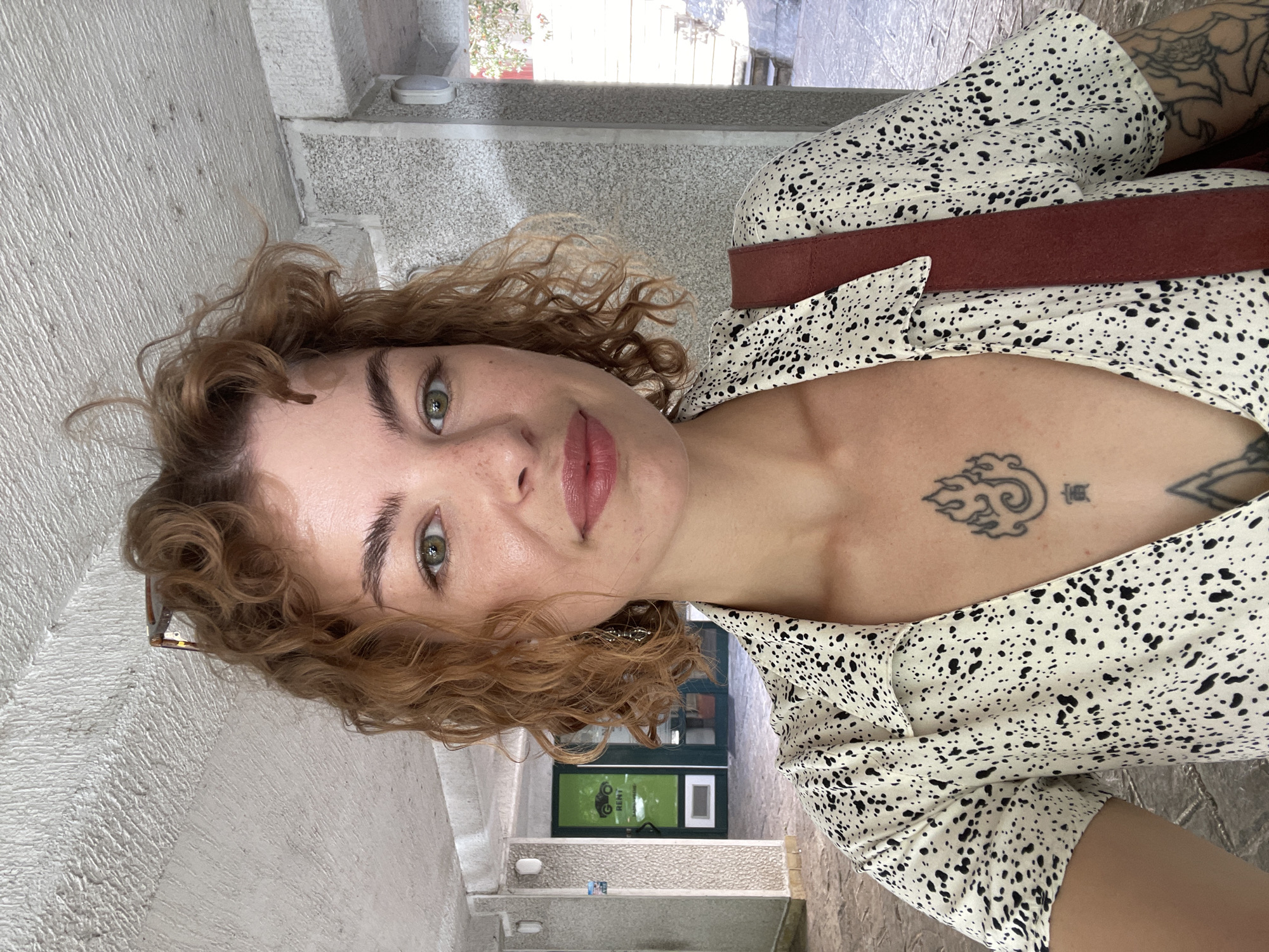 A woman with curly, reddish-brown hair and light skin is facing the camera. She has green eyes and light pink lipstick. She is wearing a white dress with black spots and a brown shoulder strap. The woman has a tattoo on her chest and another on her upper arm.
The background shows an outdoor setting that is identified as Budva, Montenegro. The architecture suggests a narrow street or alley. A green sign with "RENT" visible is partly visible in the background. The building is of light-colored stone with visible texture.