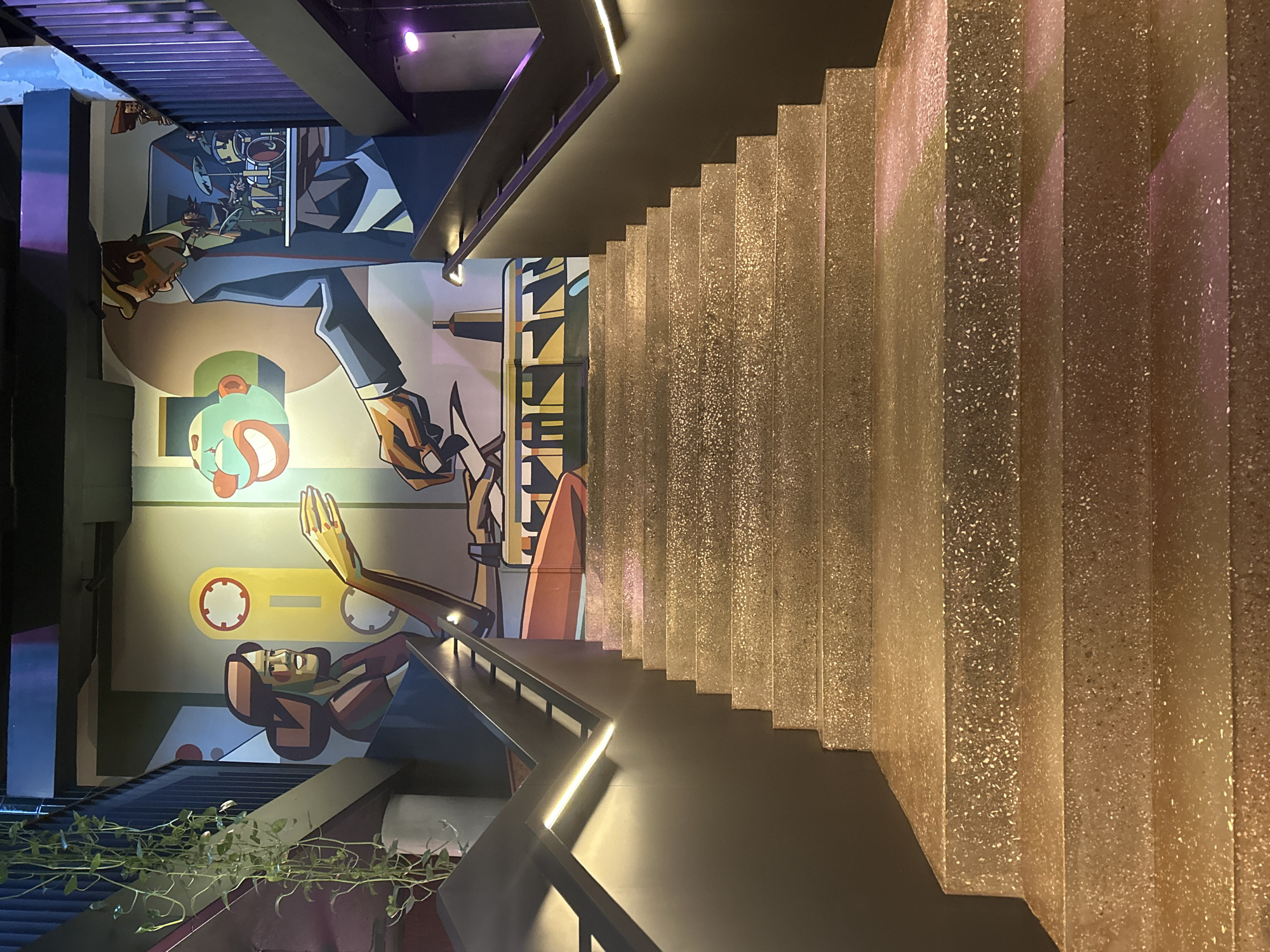 An interior shot depicts a wide staircase with a textured, speckled surface, likely terrazzo, ascending towards a mural on the wall above. The mural features a stylized, graphic art style depicting several figures. On the left, a woman with dark hair and a brown top is shown from the chest up, smiling with her hand extended. To her right, a man in a suit is depicted, holding a tray with a drum set. To the far right of the mural, three figures are seen playing drums. A clown-like face with a wide grin is also part of the mural. The staircase is flanked by dark grey railings with integrated LED lighting, casting a warm glow on the steps. Hanging plants are visible on the left side, near the top of the staircase and mural. The environment appears to be a public indoor space, possibly a restaurant, bar, or entertainment venue, given the artistic mural and modern architectural elements. The lighting is a mix of ambient interior light and the accent lighting from the staircase.