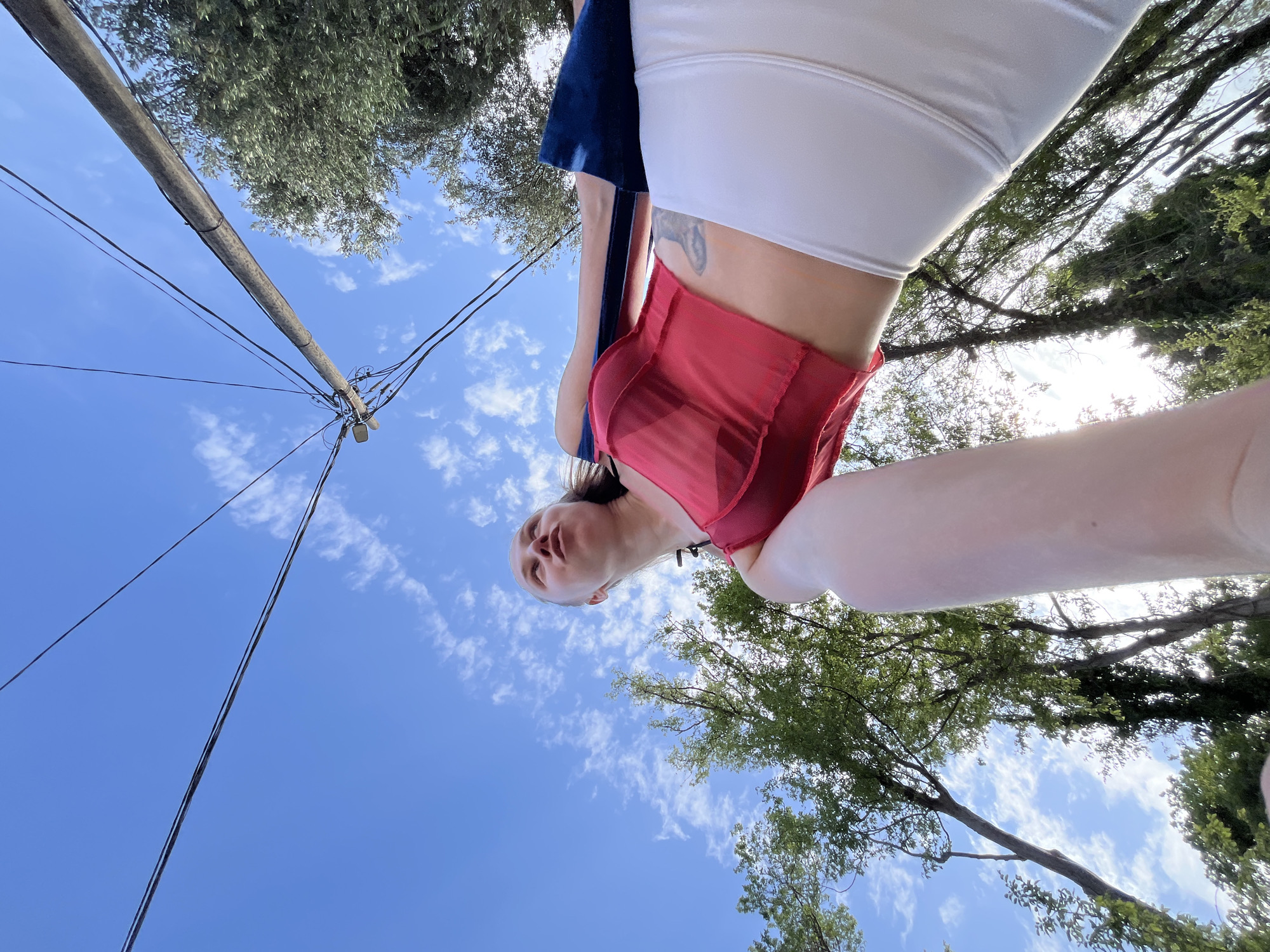 A low-angle view taken from the ground in Boreti, Montenegro, looking up at a woman with fair skin wearing a red tank top and white shorts. Her body is partially visible, and she appears to be holding the camera for a selfie. A section of blue fabric hangs near her. Behind the woman is a view of the sky, partially obscured by the branches and leaves of trees. Power lines run across the sky towards a utility pole on the left side of the frame. The sky is blue with scattered clouds.