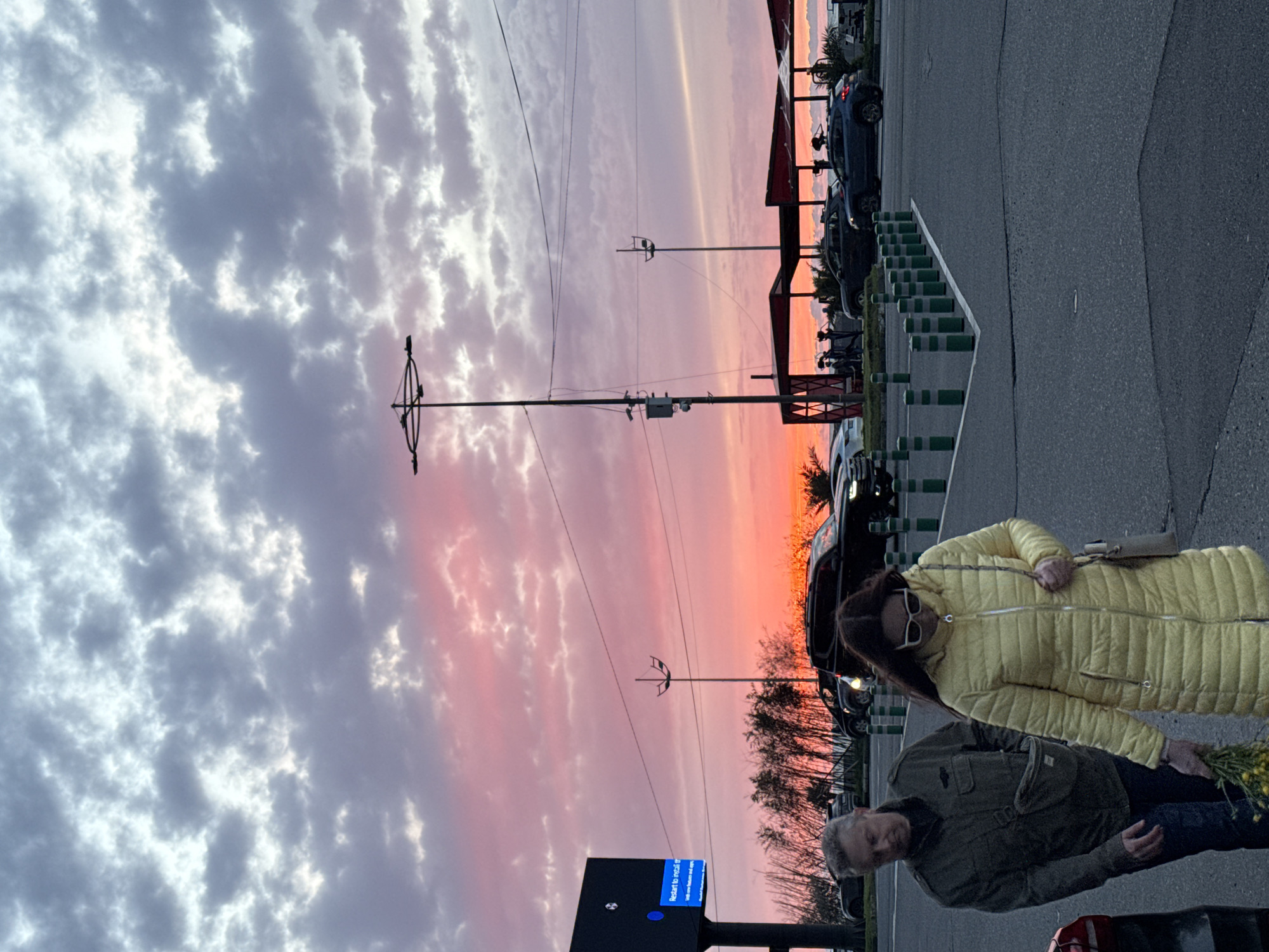 The image depicts a street scene under a sunset or sunrise sky with colorful clouds in Batumi, Georgia. An older man in a green jacket and an older woman in a yellow coat are standing near a street. The man is holding yellow flowers. Cars are parked on the side of the street, and there is signage visible. A canopy structure and street poles are also present.