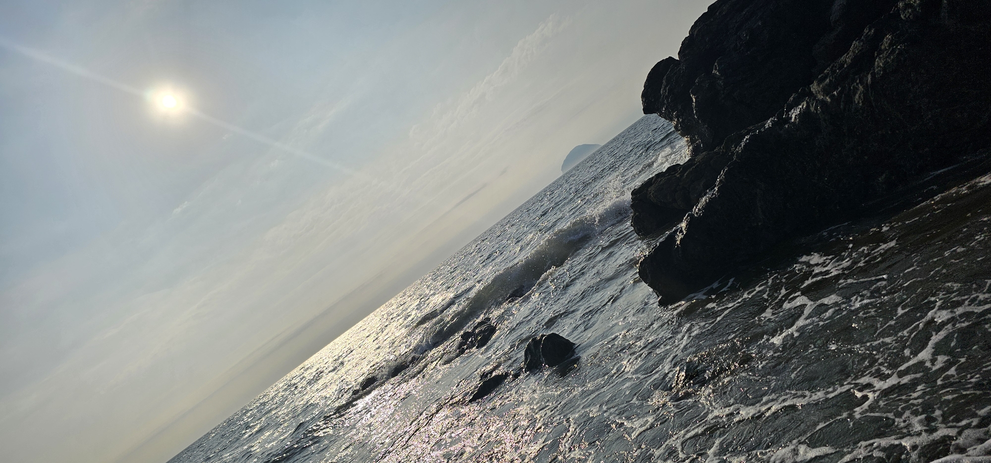 The image, taken from the coast of Girvan, United Kingdom, depicts a sunlit ocean with rocky outcrops and a distant island. The sun, a bright disc with a distinct lens flare, is positioned in the upper left quadrant, casting a shimmering path across the water. The sea itself is choppy, with waves breaking against dark, angular rocks that dominate the right side of the frame. In the distance, a small, rounded island is visible on the horizon, partially obscured by the haze. The sky above is a pale blue, streaked with wispy clouds. The overall impression is one of a rugged coastal landscape bathed in the glow of the sun.