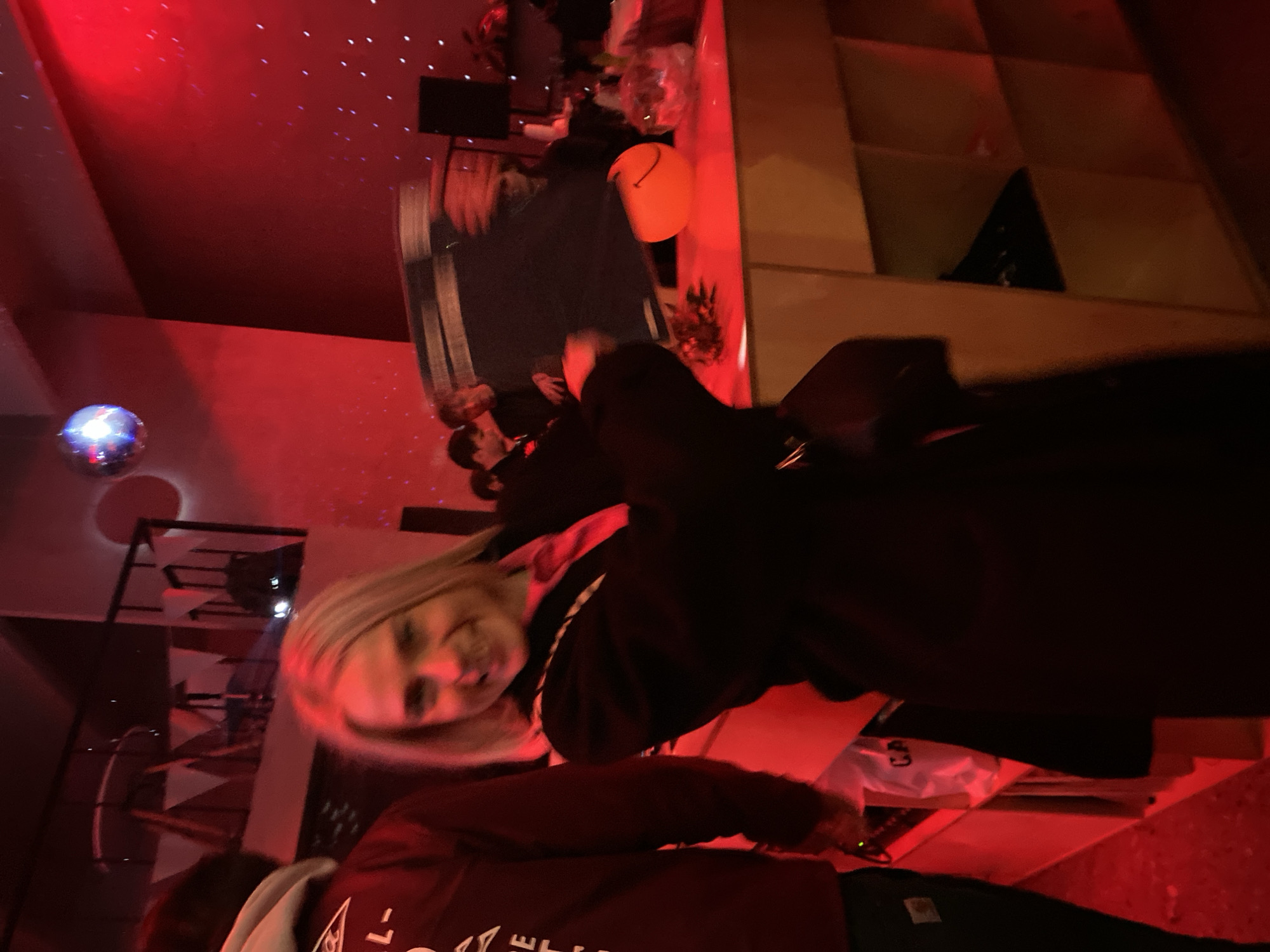 The image shows a group of people at what appears to be a party or celebration, judging by the presence of balloons and a disco ball. The lighting is predominantly red. The photo appears to have been taken in an indoor setting with a patterned wall or shelf structure in the background. No specific details indicating the location as Moscow, Russia, are visible.