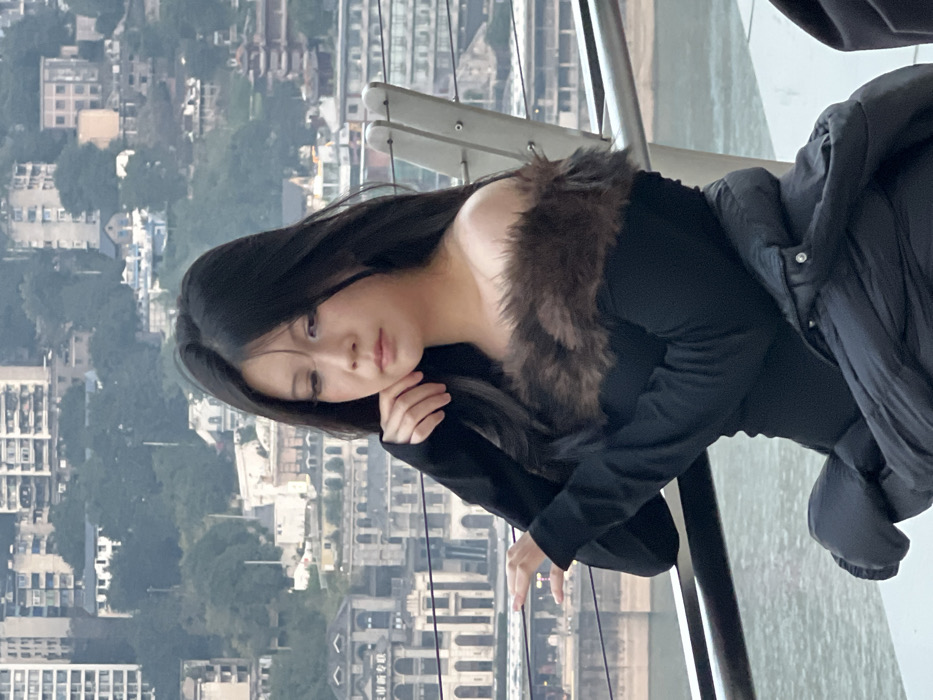 The image displays a young East Asian female in a three-quarter portrait, positioned outdoors with a city and river vista behind her. She has long, dark, straight hair, parted centrally, and is observed looking downwards with a solemn expression. Her left hand is raised, with fingertips lightly touching her jawline. She is dressed in a black, off-the-shoulder garment featuring long sleeves and a prominent dark brown faux-fur trim across the shoulders. A black puffer jacket is visible, draped across her lap in the lower right portion of the frame. In the immediate foreground, a metallic railing composed of light-colored vertical supports and dark horizontal bars separates the subject from the background. The background reveals a dense cityscape, characteristic of Yuzhong District, Chongqing, China, featuring numerous residential and commercial buildings of various heights on sloping terrain, partially obscured by atmospheric haze. Below the distant urban structures, a wide body of water, indicative of a major river, flows horizontally across the mid-ground. The scene is illuminated by diffused natural light, suggesting an overcast day or late afternoon.