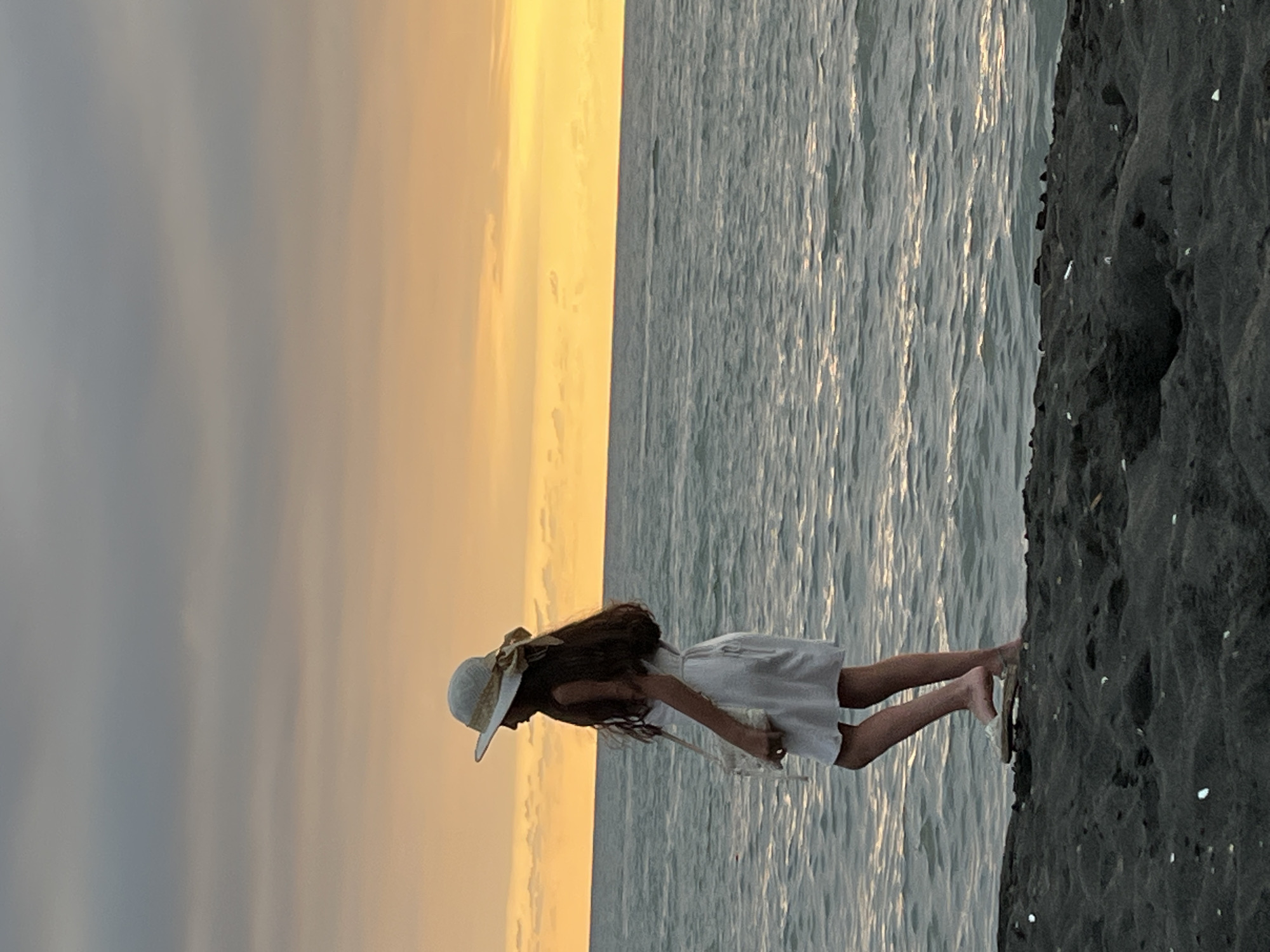 A young person, wearing a white dress and a wide-brimmed straw hat, stands on a black sand beach in Ureki, Georgia. The person is facing right, with their body in profile, and their long, dark hair is visible. They are holding a small, light-colored bag in their left hand. The sea stretches out before them, with gentle waves reflecting the warm glow of a sunset. The sky above is a gradient of orange and grey clouds. The beach appears to be made of dark, coarse sand with a few scattered small, light-colored objects.