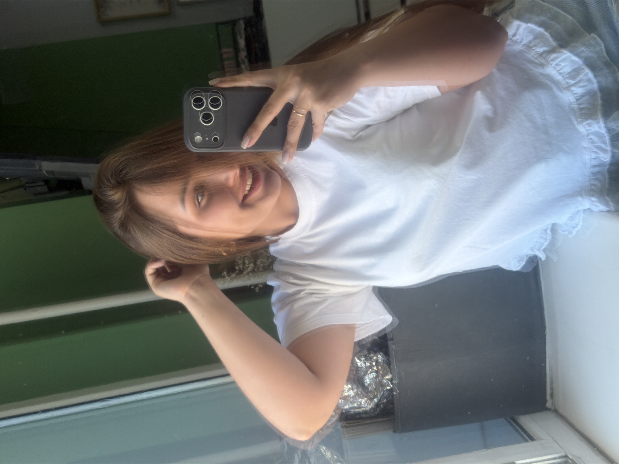 A young woman with brown hair and a white t-shirt is taking a selfie in front of a window. She is holding a grey smartphone with three cameras. The location appears to be indoors, with a view of what could be a green-toned exterior. The presence of window frames suggests a residential setting, but without further visual cues it's difficult to conclusively determine it is located in the city of Chaikovsky, Russia.