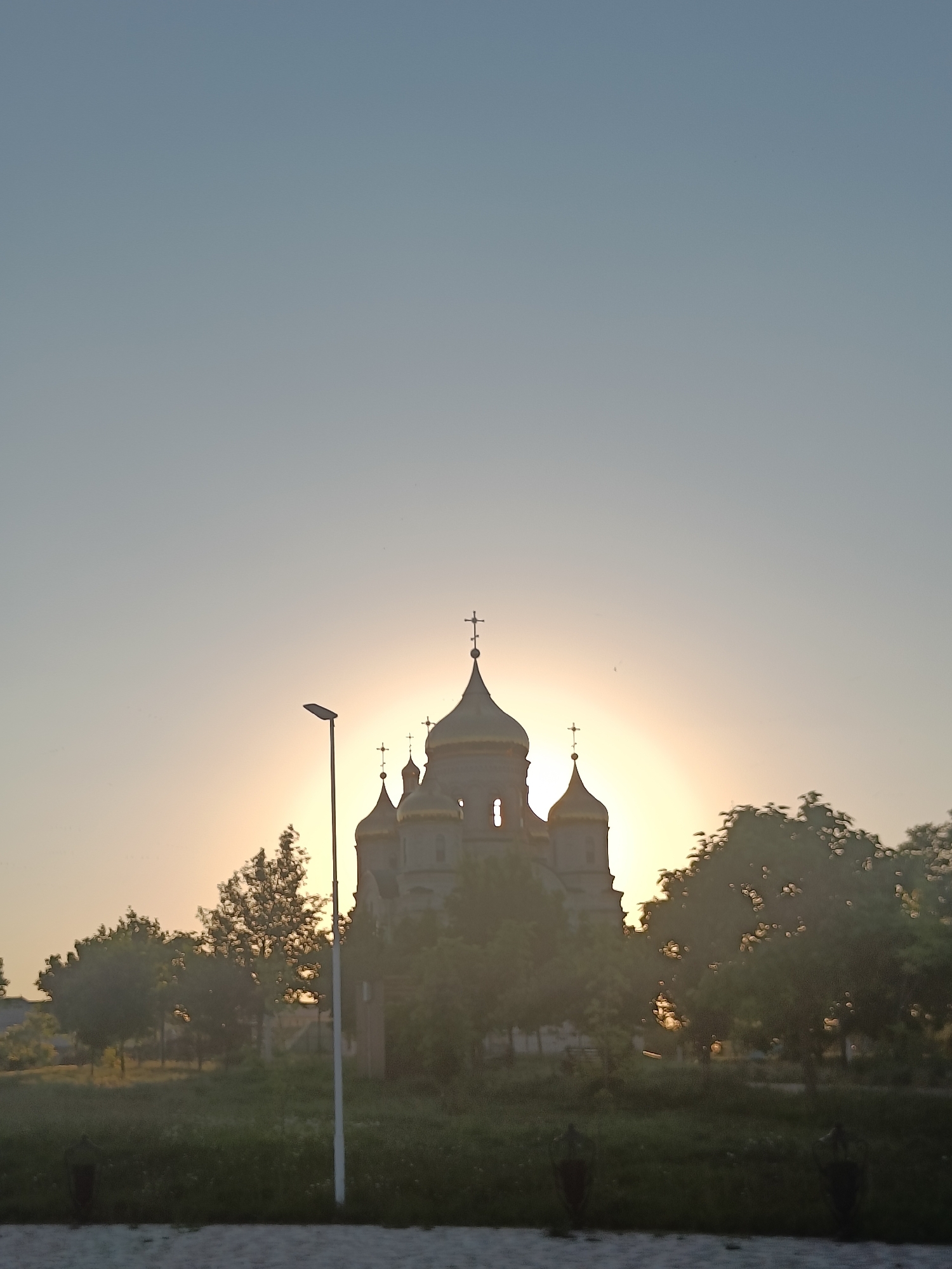 The image depicts a view of the Church of the Transfiguration in Aleksandrovskoye, Russia, during sunset. The sun is partially obscured by the church's domes, creating a bright halo effect. Several golden domes, each topped with a cross, are visible on the church structure, which appears to be made of concrete or stone. In the foreground, a tall, slender street lamp stands to the left of the church. Lush green grass and trees surround the church, with their leaves also illuminated by the setting sun, creating a dappled light effect. The sky is clear and transitions from a light blue at the top to a soft orange near the horizon. The overall scene is tranquil and serene, with the warm glow of the sunset casting a peaceful ambiance over the religious building and its surroundings.