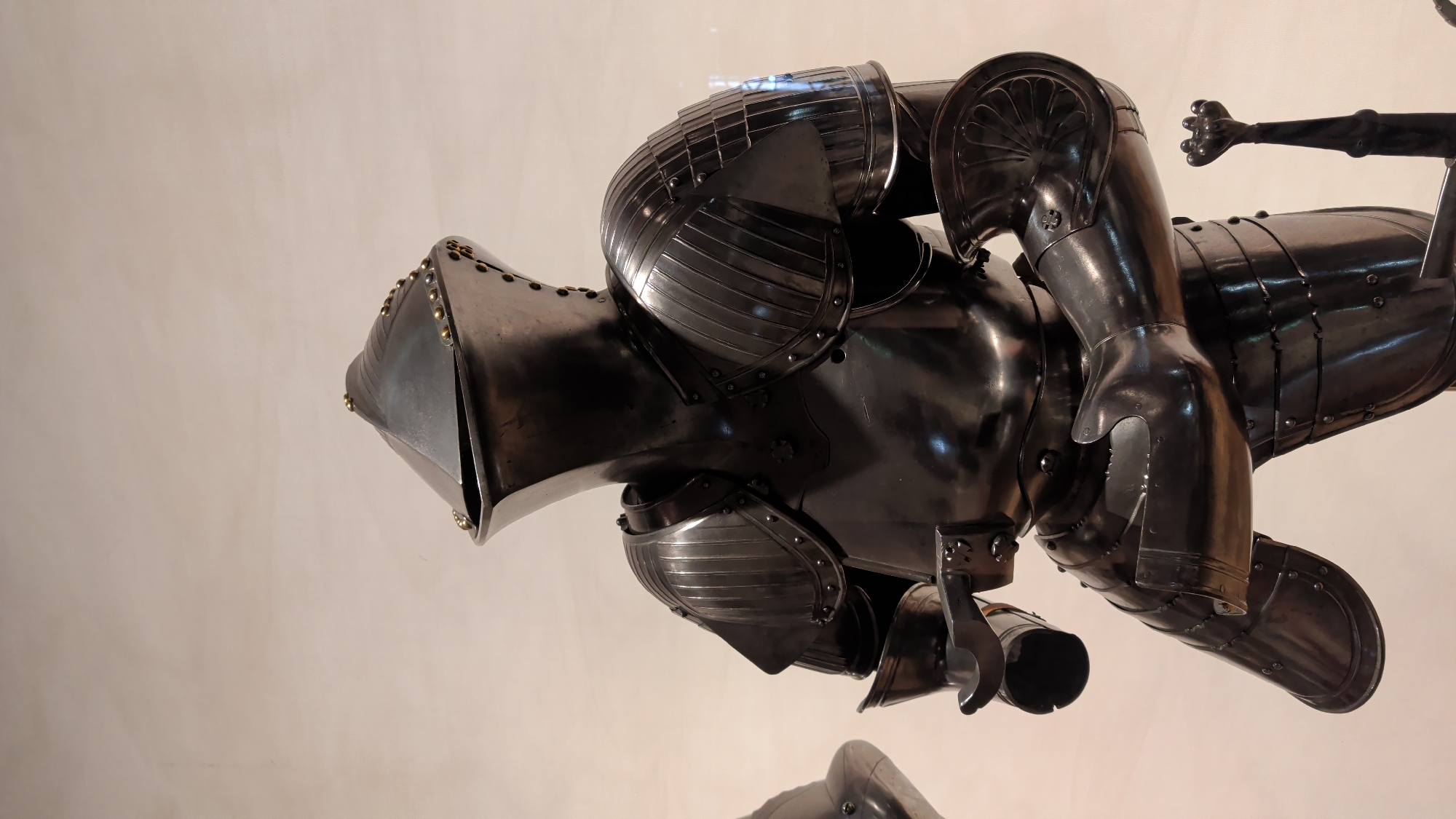 A full suit of polished black metal armor, likely from the 16th century, stands upright against a neutral, light-colored background. The armor covers the entire body, from the conical helmet with a hinged visor to the armored boots. The armor features segmented shoulder pieces, a breastplate, vambraces, and gauntlets. A decorative sword hilt is visible to the lower right of the frame. The overall appearance is that of a museum exhibit. The location is the Royal Castle in Warsaw, Poland.