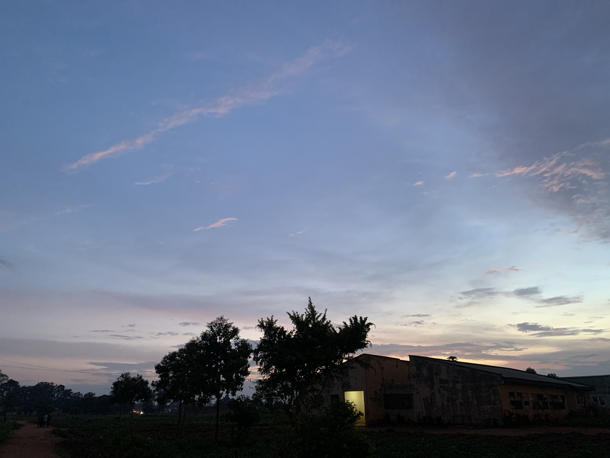 The image captures a twilight scene in Kuru Karama, Nigeria, with the sky transitioning from a soft blue to hues of pink and orange as clouds drift across. In the foreground, silhouetted against the darkening sky, are several trees. To the right, a weathered, light-colored building with a sloped roof is visible, its facade showing signs of age. A bright, yellow light emanates from an open doorway on the building. In the distance, a dirt path leads towards a group of indistinct figures, and the silhouettes of more trees suggest a rural or semi-rural environment. The overall impression is one of dusk settling over a quiet area.