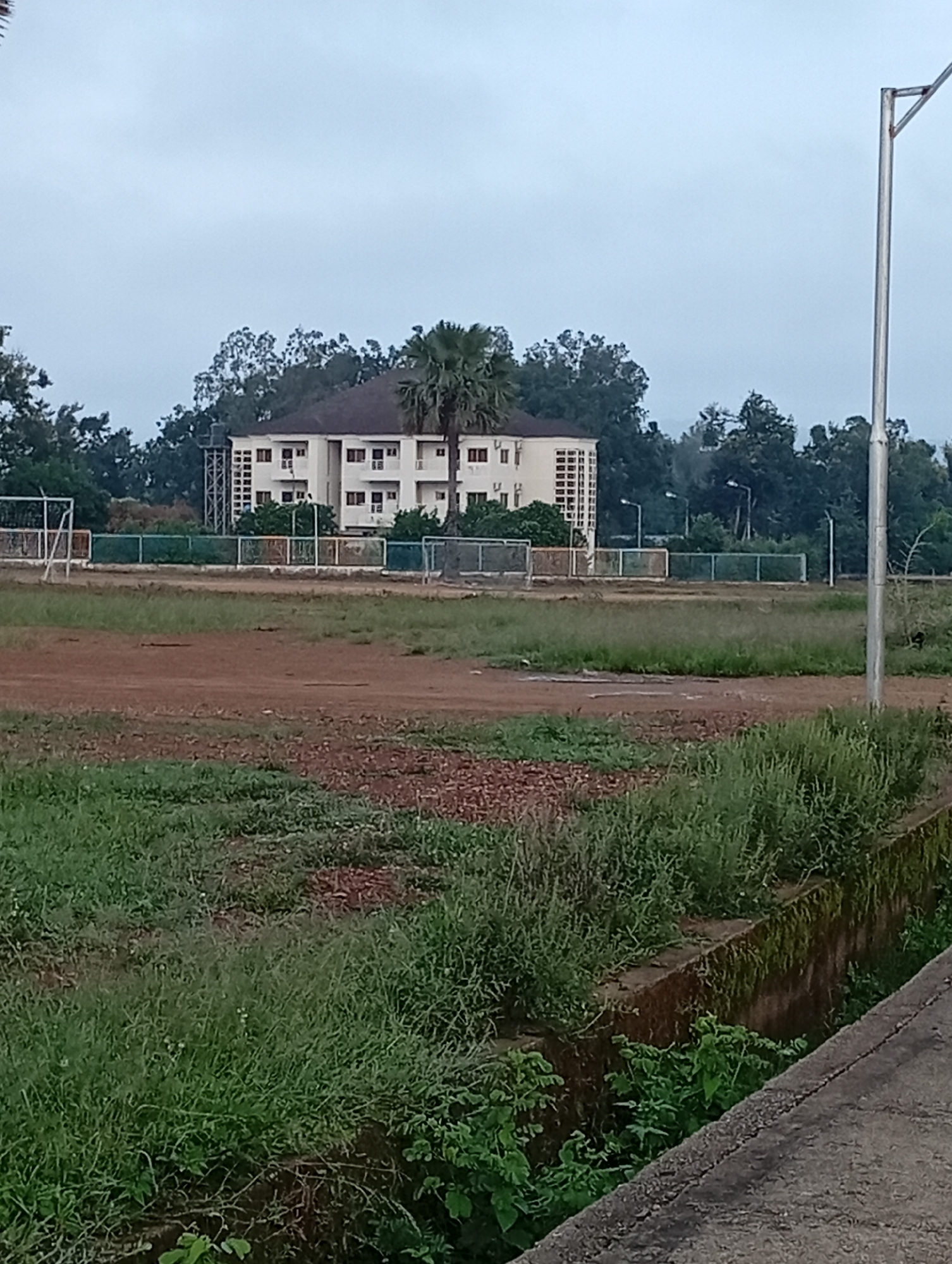 The image depicts an outdoor scene, likely a sports field or training ground, with a large, cream-colored building in the background. The building appears to be multi-story with numerous windows. In front of the building, a fence encloses an area with what looks like a sports field, possibly for soccer, indicated by the presence of a goalpost. Trees, including a prominent palm tree, surround the building and extend into the background. The foreground of the image shows a dirt and grassy field, with some patchy brown earth and greener grass. A concrete structure, possibly a drain or retaining wall, runs along the right side of the foreground, covered in moss and vegetation. A tall, silver light pole is visible on the far right side of the image. The sky is overcast. The scene suggests a somewhat neglected or developing area, possibly part of a school, university, or sports complex in Jos, Nigeria, given the environmental context and architectural style. There are no people visible in the image.