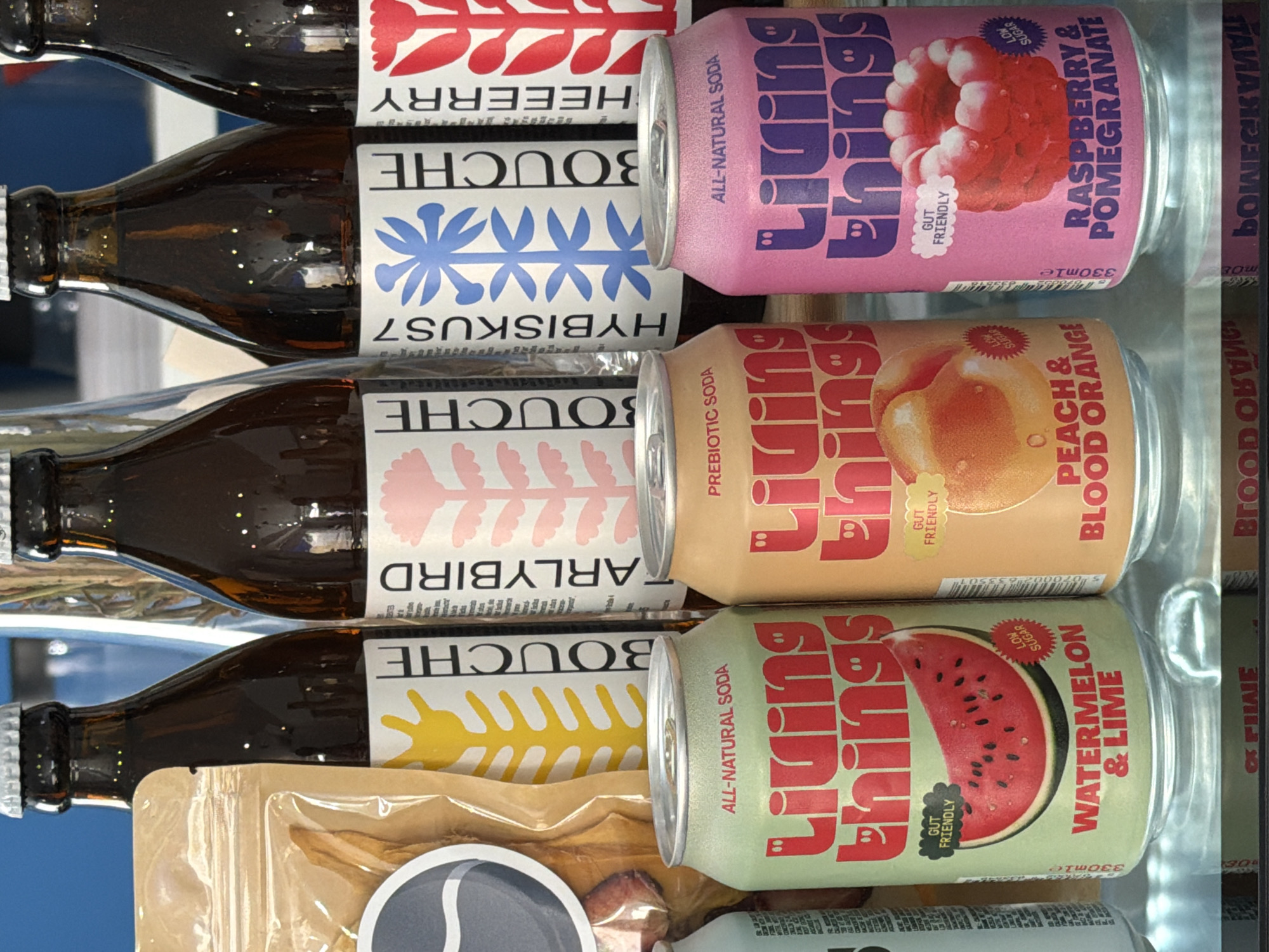 The image depicts a shelf display of beverages and snacks, likely in a retail setting. In the foreground, three colorful aluminum cans of "Living Things" soda are prominently displayed. The can on the left is green and features an illustration of a watermelon slice, with text indicating "WATERMELON & LIME," "ALL-NATURAL SODA," "GUT FRIENDLY," and "LOW SUGAR." The middle can is a peachy-orange color with an illustration of a peach or apricot, labeled "PEACH & BLOOD ORANGE" and "PREBIOTIC SODA." The can on the right is pink, with an image of a raspberry-like dessert, and reads "RASPBERRY & POMEGRANATE," "ALL-NATURAL SODA," "GUT FRIENDLY," and "LOW SUGAR."
Behind the cans, several brown glass bottles with white labels are visible. These bottles appear to be beer or some other bottled beverage. The labels have varying graphic designs and text, including "BOUCHE," "EARLYBIRD," "HYBISKUS7," and "HEEERRY," each accompanied by stylized illustrations.
To the far left, a translucent plastic bag filled with dried fruit or jerky is partially visible. The overall environment appears to be well-lit, possibly a shop counter or a refrigerated display case, suggesting a commercial space.
This scene is observed in Valencia, Spain, within a store called "La Finestra" located in the Ruzafa neighborhood. The establishment specializes in natural and organic products, including a selection of craft beers, sodas, and artisanal food items. The display is arranged to highlight the vibrant branding and natural ingredients of the "Living Things" sodas.