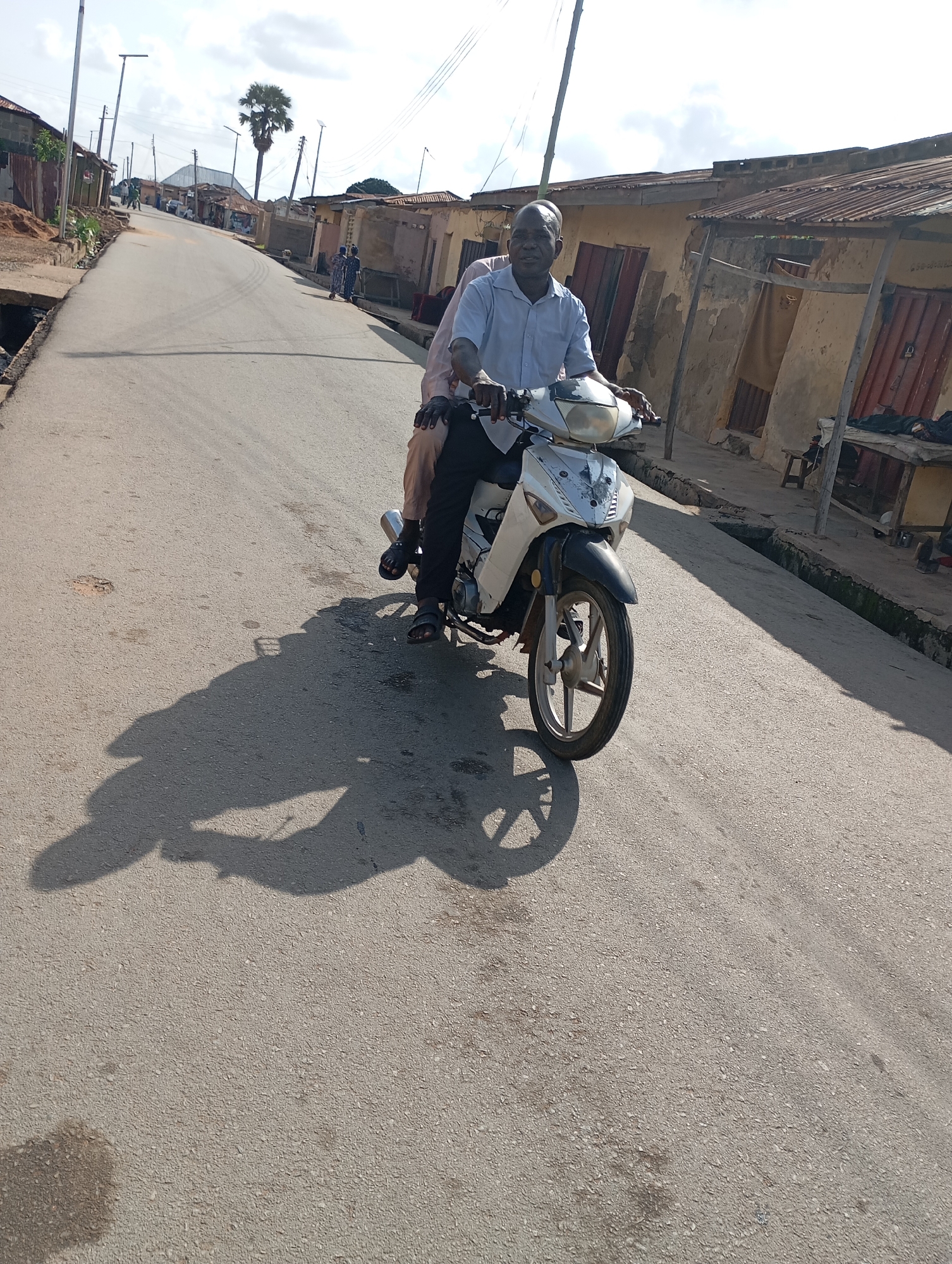 A man and a woman are riding a white and black motorcycle down a paved road in what appears to be a village or small town in Shendam, Nigeria. The man is driving, wearing a light blue collared shirt and dark pants. The woman is a passenger behind him, her arms wrapped around his waist. They are both wearing sandals. The motorcycle is positioned in the foreground, casting a long shadow on the road.
The scene is lined with simple buildings, many with corrugated metal roofs and weathered walls, suggesting a rural or semi-urban setting. Some buildings have open doorways. In the distance, a tall palm tree stands out against a bright, partly cloudy sky. Streetlights are visible along the road, indicating some infrastructure. The road surface is asphalt and appears to be in decent condition, with some minor debris and water stains.
The sun is high in the sky, suggesting it is daytime, likely mid-morning or early afternoon, given the strong shadows. The weather appears to be warm and dry. The overall mood seems neutral, with the focus on the mode of transportation and the surroundings. There is no visible text in the image. The activity is simply traveling by motorcycle through the village.