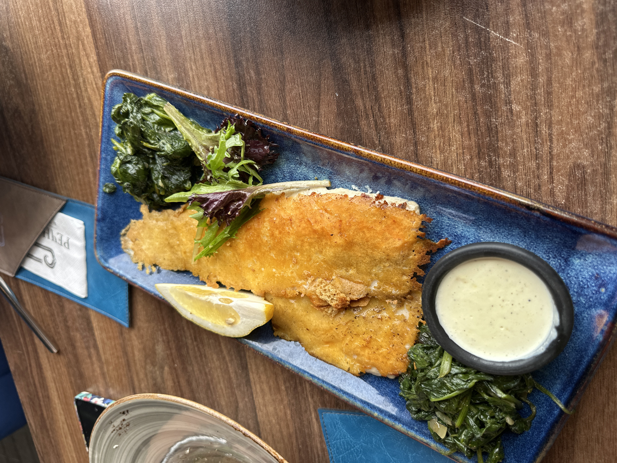 A plate of food is placed on a wooden table. The rectangular blue plate features fried fish, a lemon wedge, spinach, salad greens, and a small black bowl of white sauce. The table is brown. A blue and white menu or coaster is visible to the left of the plate. A partially visible glass bowl is present in the lower left corner. The photograph was likely taken in Vidin, Bulgaria.