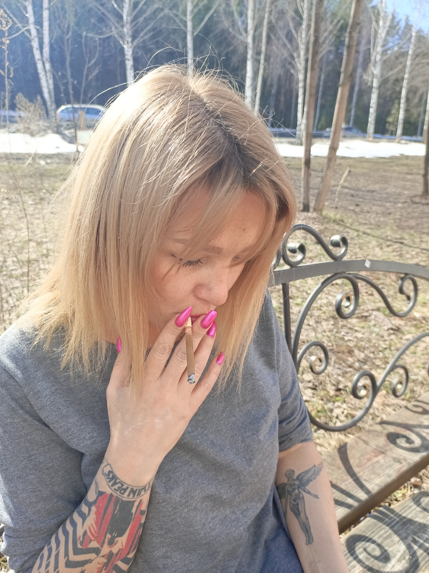 A woman with blonde hair and pink manicured nails sits on a wrought iron bench in what appears to be a park or wooded area. She is holding a lit cigarette or cigarillo in her right hand, near her mouth. She is looking down.
She is wearing a gray t-shirt, and tattoos are visible on both forearms. The tattoo on her left forearm includes red, black, and gray colors, and the phrase "Twin Peaks" in a circular banner. The tattoo on her right forearm is of an angel.
In the background, there are birch trees, and patches of snow on the ground, suggesting it is early spring. The bright sunlight indicates a clear day.
While the description states the image contains a location in the city of Chaikovsky, Russia, there is no visible information in the image to confirm this statement.
