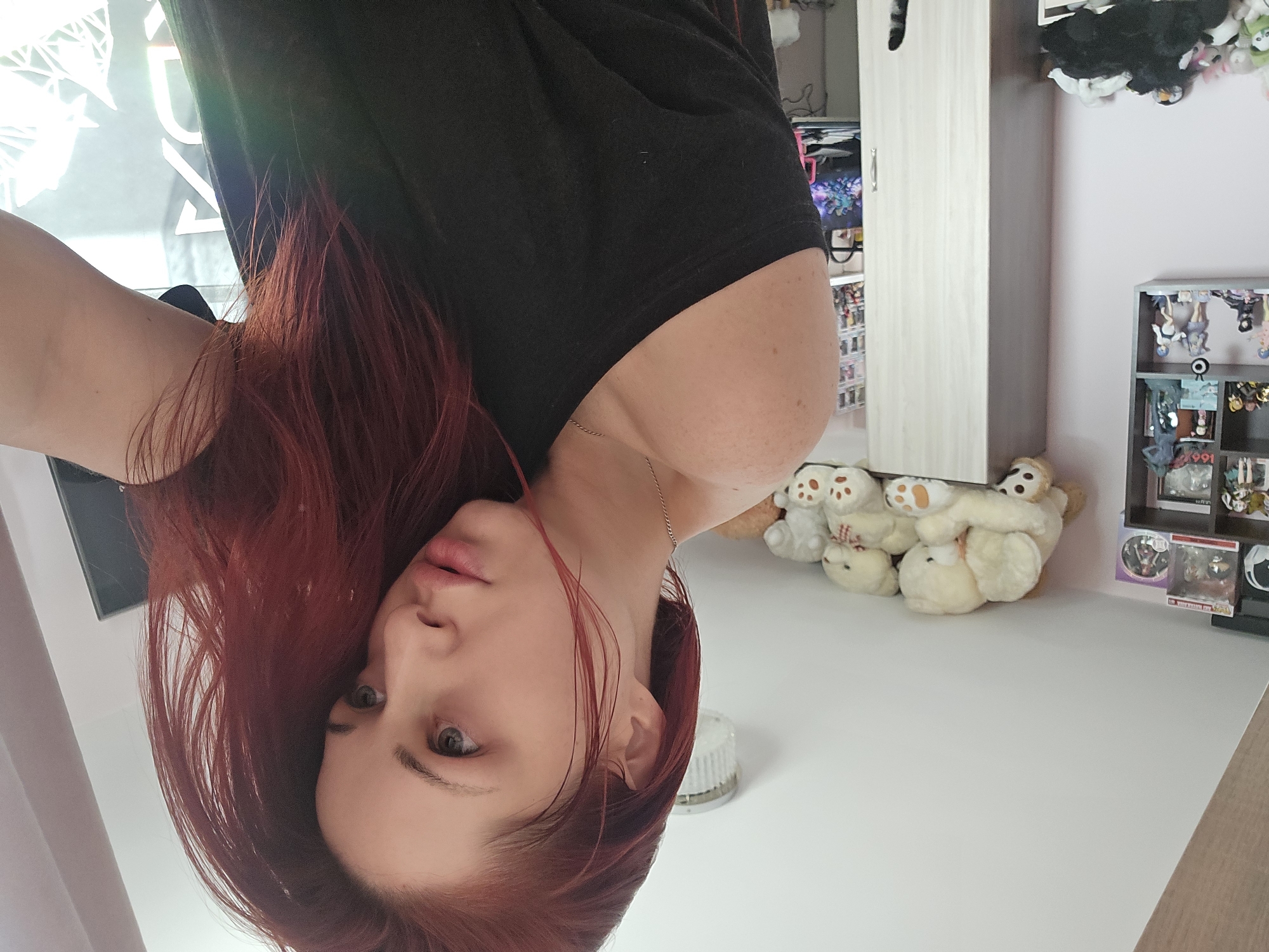 A young woman with red hair, wearing a black top, is looking at the camera. She is positioned upside down relative to the background.
The background includes:
* A light pink wall.
* A white cabinet.
* Stuffed animals, including teddy bears, arranged near the cabinet.
* A display shelf with various figurines and objects.
* A white surface, possibly a table or desk.
* A light fixture on the white surface.
* A window with light filtering in.
There is no evidence in the image to suggest it was taken in Burgas, Bulgaria.