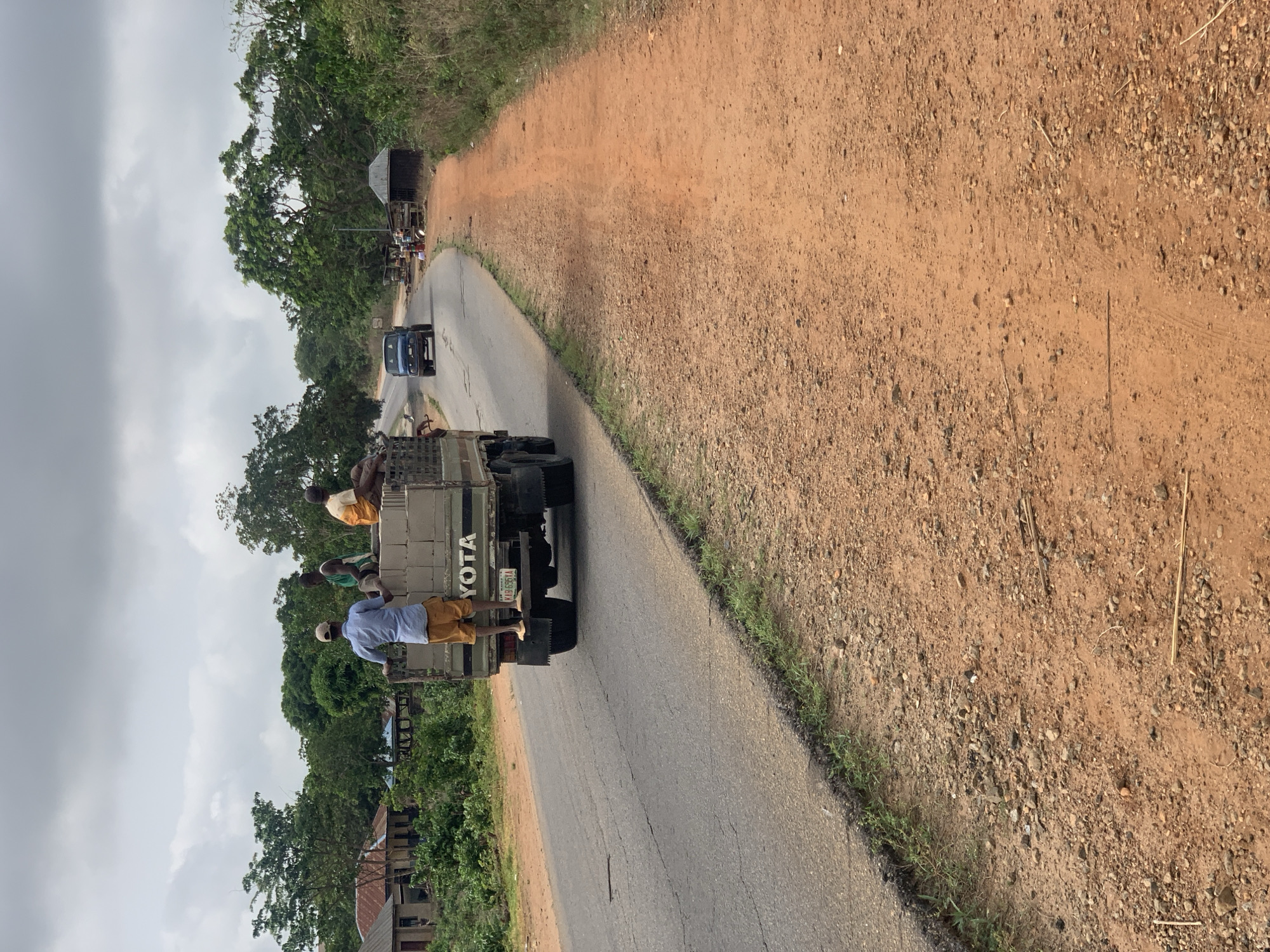 The image depicts a road in Kabba, Nigeria. A light-colored Toyota truck is traveling along the road, carrying multiple people in its bed. A blue vehicle and a larger truck are further up the road. The surrounding environment includes trees, vegetation, and dirt on the road's shoulder. The sky is overcast. Houses are visible in the background. The license plate of the truck reads "KAB 685 KA".