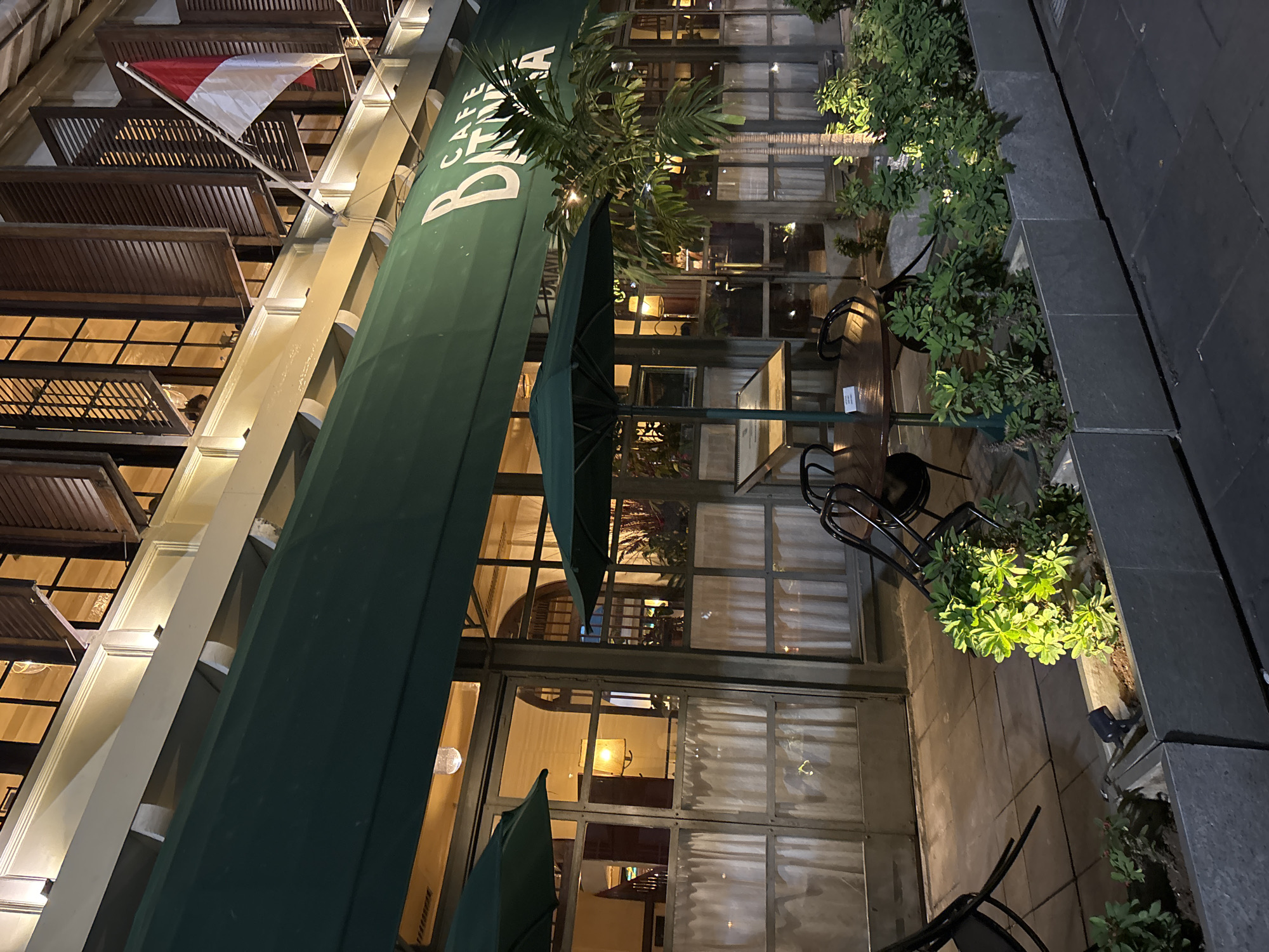 The image shows the facade of "Cafe Batin" located in Kota Tua, West Jakarta, Indonesia. The cafe has a green awning with white lettering spelling out "CAFE BANTEN". An Indonesian flag flies from a flagpole on the upper story. The building features large windows with wooden shutters and is illuminated by interior lighting. Outside the cafe, there are tables and chairs, some of which are tilted. Planters with green foliage are positioned along the sidewalk. The scene is depicted at night, with artificial lighting illuminating the cafe and its surroundings.