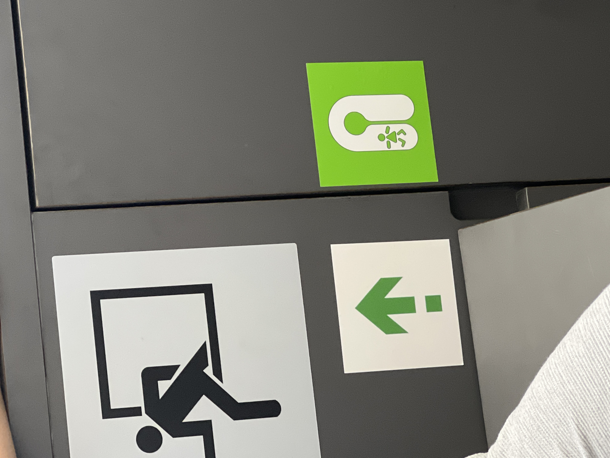The image shows a close-up of a dark gray surface, likely a wall or door, with three signs affixed to it. On the left, a white square sign with a black outline depicts a stylized figure exiting a black rectangular frame, indicating an exit. Below this, another white square sign displays a green upward-pointing arrow with a smaller green square at its base. To the right, a bright green square sign features a white graphic of a stylized person within an arch shape. This could indicate a family restroom or a changing facility. The lighting suggests an indoor environment. There is no visible context to determine its specific location within the city of Vilnius, Lithuania.