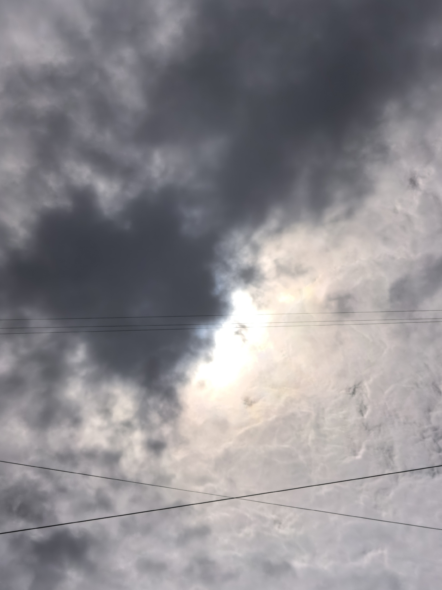 The image shows a cloudy sky with the sun peeking through the clouds. The sun is off-center, to the right of the frame, creating a bright halo. There are several dark, heavy clouds obscuring parts of the sky, and lighter, wispy clouds also visible. In the foreground, there are three horizontal dark lines that appear to be power lines. The perspective suggests the photo was taken looking upwards. The image does not contain any people or specific identifiable landmarks that would place it in the city of Krasnoyarsk, Russia.