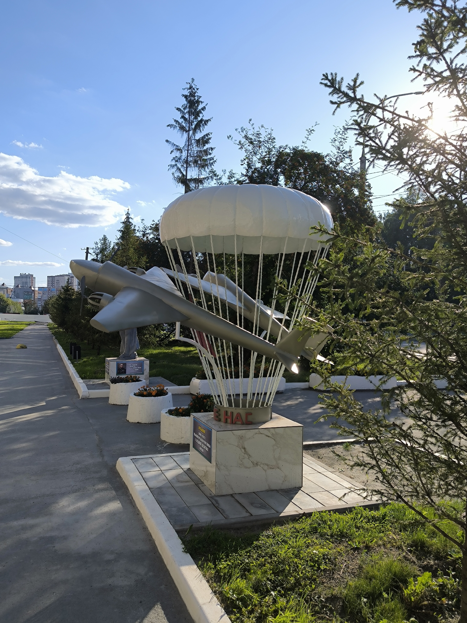 An outdoor monument, likely located in Novosibirsk, Russia, depicts an airplane suspended beneath a parachute. The monument is situated on a paved pathway, bordered by a concrete curb and grass. In the background, several apartment buildings and trees are visible under a clear blue sky with scattered clouds. The sun is shining brightly, casting shadows and creating a lens flare effect through the branches of a nearby evergreen tree in the foreground. The monument itself features a silver-colored airplane model, a white parachute canopy, and white supports connecting the two. The base of the monument is a marble-like structure with inscribed text. Several white planters filled with orange flowers are placed along the pathway in front of the monument.
