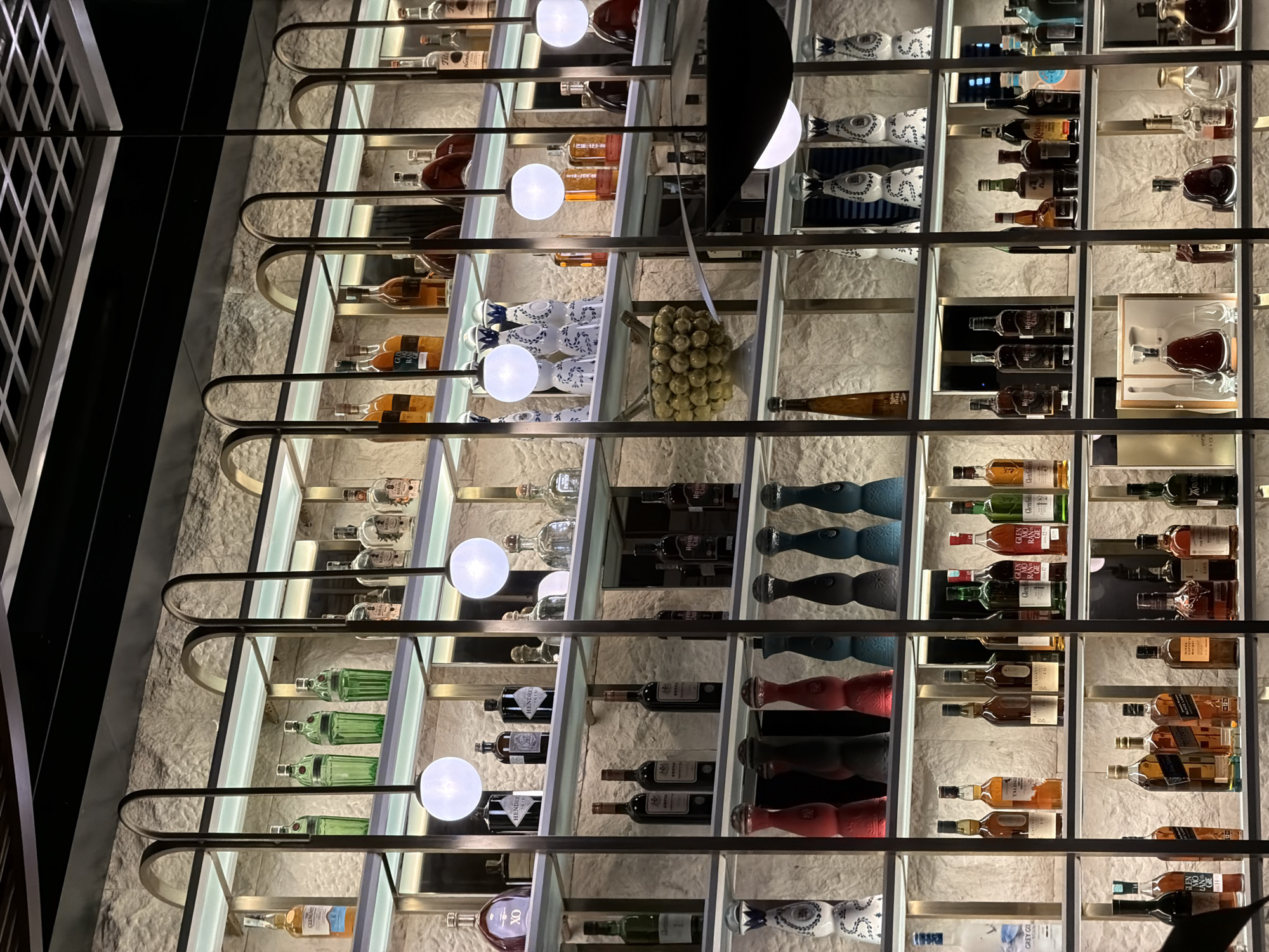 Here is a detailed description of the image:
**Scene:** The image showcases a well-lit liquor display case. It features a diverse selection of alcoholic beverages, including various types of liquor bottles arranged on multiple shelves within the case.
**Objects:** The display consists primarily of liquor bottles with different shapes, sizes, and labels. A large, modern light fixture is visible in the foreground, casting light on the display.
**Environment:** The shelves are illuminated, emphasizing the bottles and their contents. The wall behind the display appears to be made of stone or a textured material.
**Location:** While the specific city of Budva, Montenegro is not discernible from the image itself, it can be inferred based on the prompt. Therefore, the location is likely a bar, restaurant, or retail store in Budva.