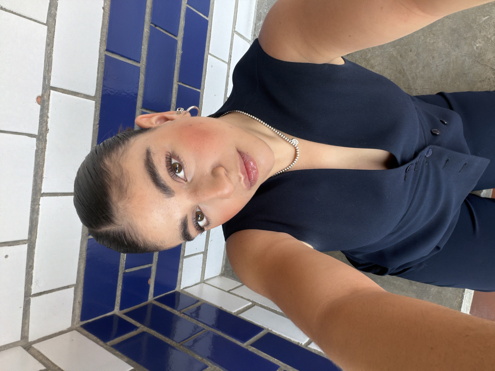 A woman is taking a selfie outdoors. She is wearing a dark blue v-neck garment and a delicate silver chain necklace. Her hair is pulled back, revealing silver earrings. The background consists of tiled surfaces with alternating white and blue tiles. The setting may be in San Bernardino Tlaxcalancingo, Mexico.