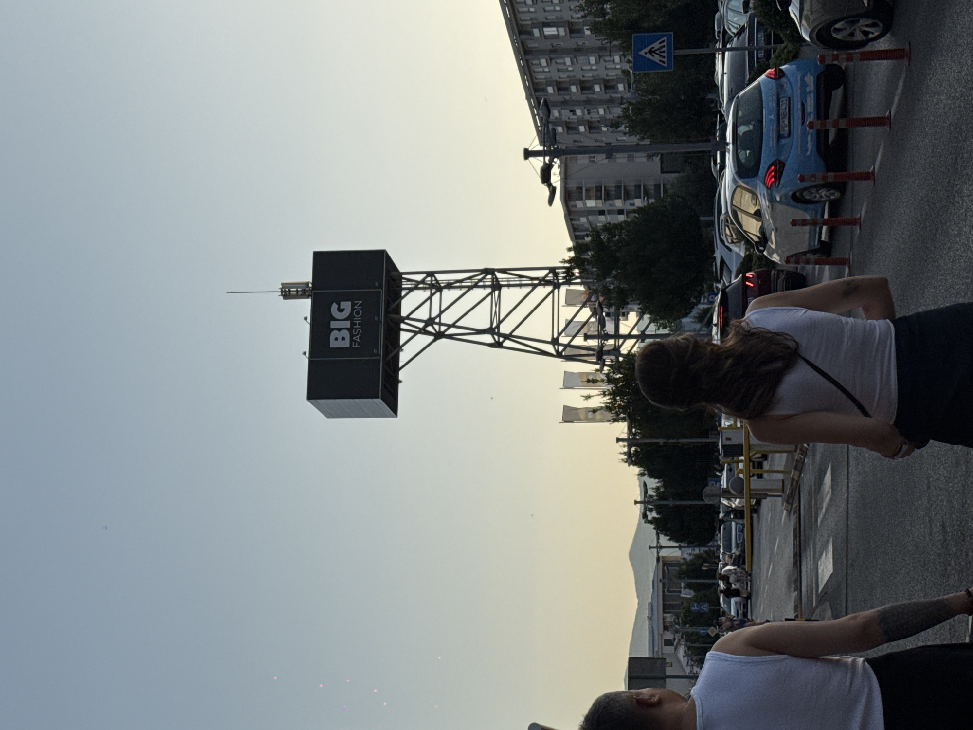 In an outdoor urban scene, two individuals, seen from behind, walk away from the camera. The person on the left wears a white tank top and black bottoms, with a tattoo visible on their left arm. The person on the right also wears a white tank top and black bottoms. In the background, a large black sign reading "BIG FASHION" is mounted on a metal tower. Several cars are parked and driving on a road, and buildings and trees are visible. The sky is clear and light blue.