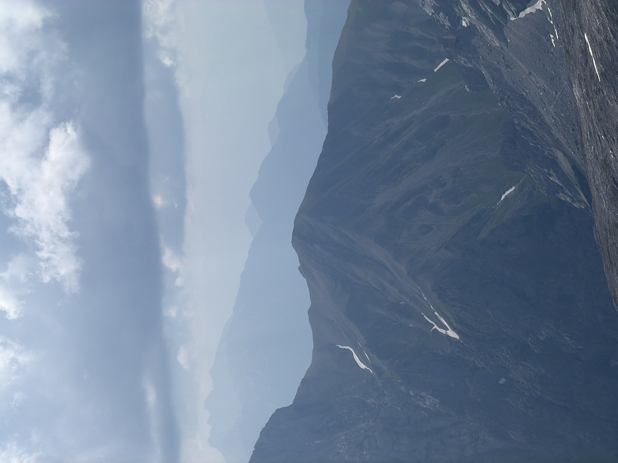 The image shows a view of layered mountain ranges under a cloudy sky. The foreground features a steep, rocky slope with patches of snow and green vegetation. Several distinct mountain ridges are visible in the background, fading into a hazy blue, suggesting distance and atmospheric perspective. The sky is dominated by heavy, grey clouds with some lighter, brighter areas, indicating diffused daylight. There are no people or man-made objects visible in the image. The scene is natural and appears to be a high-altitude mountain landscape. This view is likely of the Bavarian Alps, a region that includes Garmisch-Partenkirchen, Germany, known for its dramatic mountain scenery.