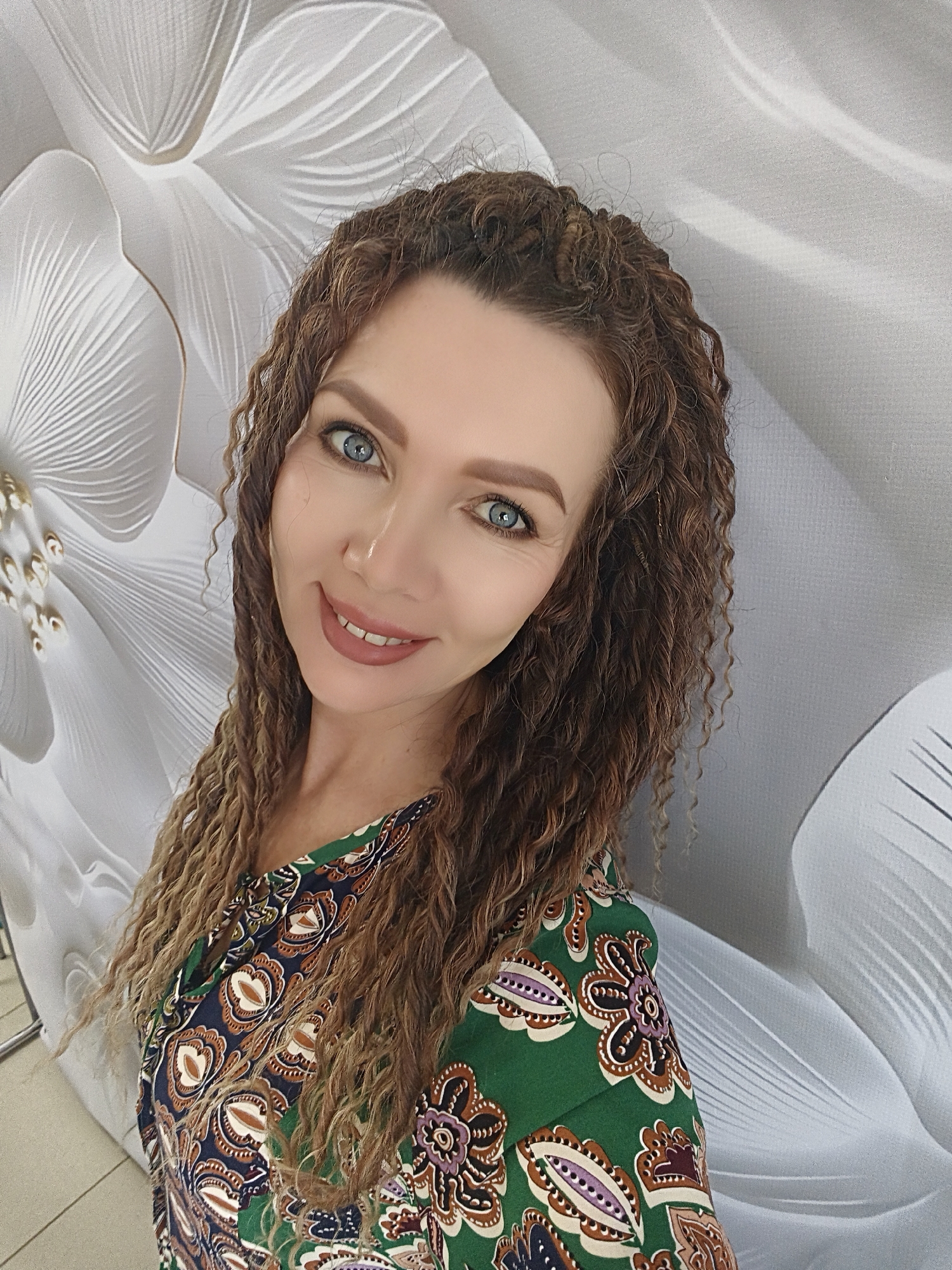 A woman with blue eyes and curly brown hair is centered in the image. She is smiling and wearing a green patterned shirt. The background is a wall with a white flower design. The location is unknown.