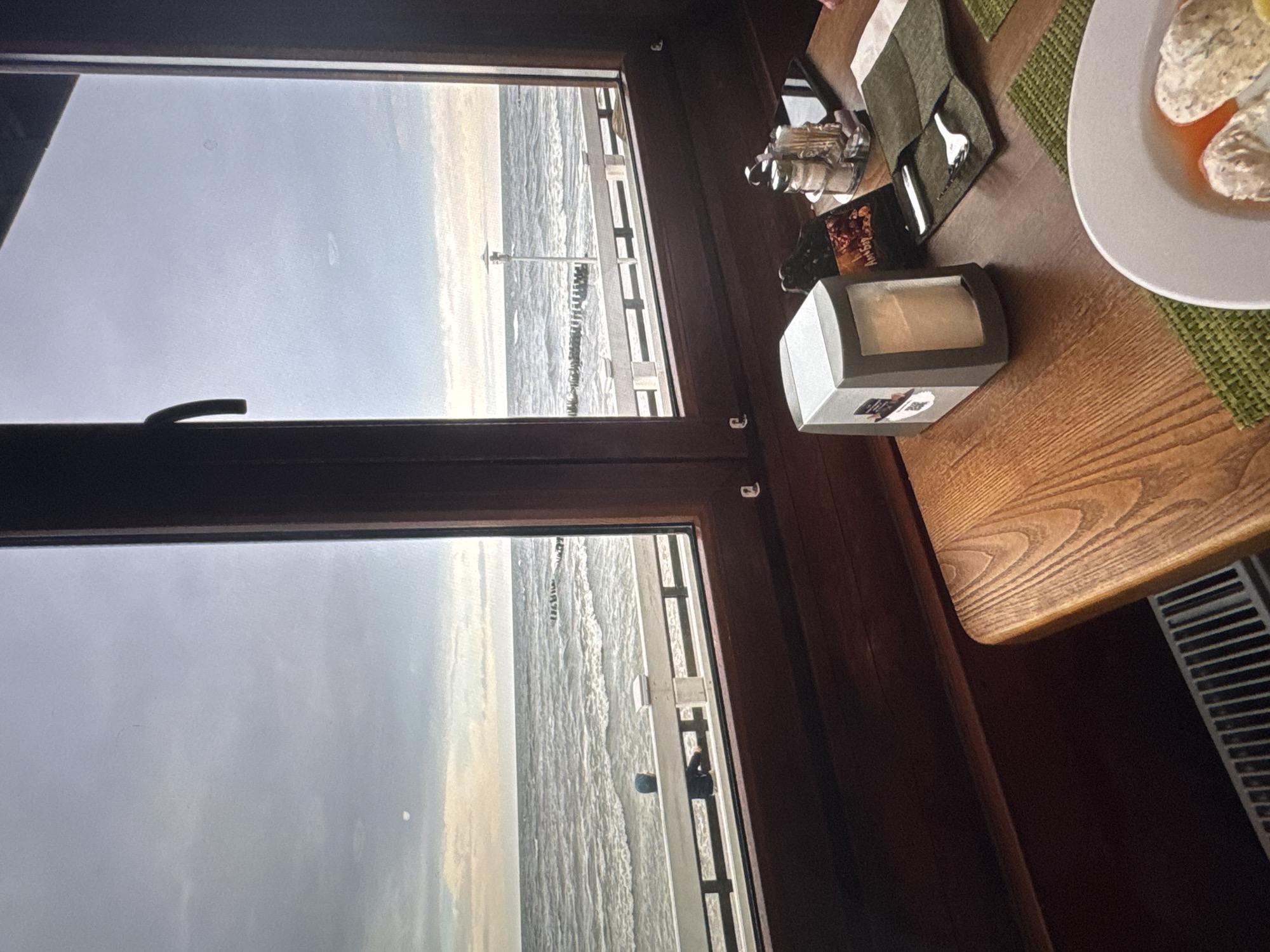 The image depicts an indoor scene with a view of the sea through a large window. The window frame is made of dark wood. Outside, the sea is choppy under a cloudy sky, with a breakwater visible in the distance. A person wearing a blue hood is leaning against a white railing on a promenade overlooking the sea.
On the table inside, a napkin dispenser, a salt and pepper shaker set, a phone, and a cutlery set are arranged. A plate with food, possibly dumplings in broth, is also visible on the table. The table itself is made of wood. A white radiator is present at the bottom left of the image. The scene appears to be a restaurant or cafe. Based on the visual elements, this location is in Новинки, Russia, a coastal town known for its seaside views.
