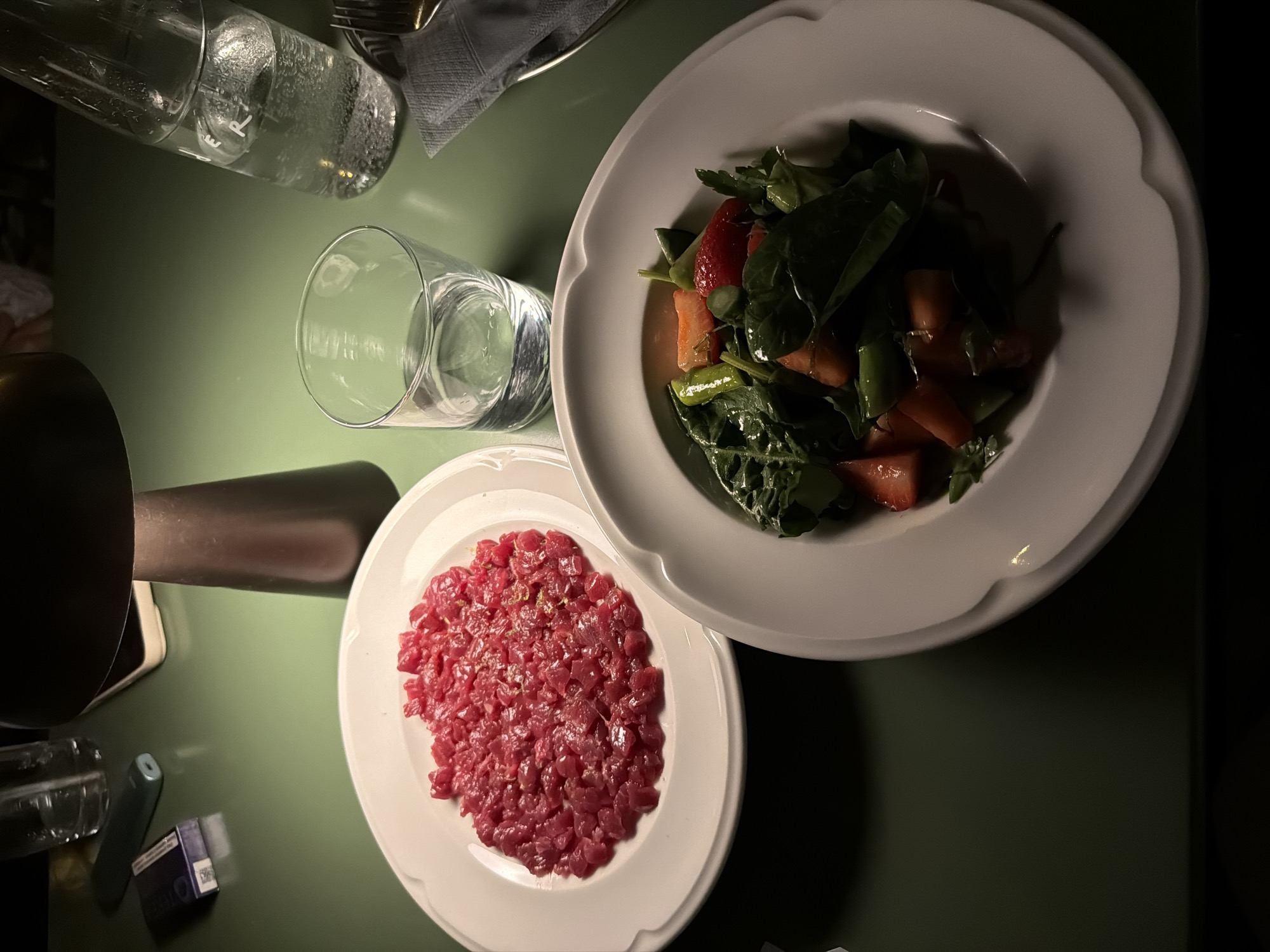 A table in a restaurant in Belgrade, Serbia, is set with two white plates, one containing steak tartare and the other a salad with greens, tomatoes, and other vegetables. Two glasses of water are also present. A silver fork is laid next to a grey napkin. Other items on the table include a black object, a small blue box, and a clear cylindrical object. The table has a green surface. The light is dim and overhead.