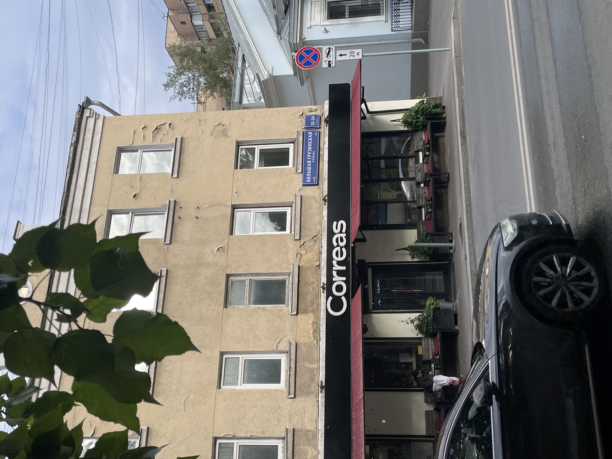 The image shows a street view in Moscow, Russia. A light brown, multi-story building with several windows lines the street. The building's facade shows signs of peeling paint. A black vertical sign with the white text "Correas" is mounted on the side of the building, next to a red awning over a storefront. The storefront appears to be a cafe or restaurant with large glass windows. Inside, a person can be seen sitting at a table.
Adjacent to the building is a paved street with white lane markings. A black car is parked on the side of the street. Traffic signs are visible on a pole. One sign indicates a parking restriction, and another shows an arrow pointing upwards with "20 M" next to it, likely indicating a distance.
A tree with green leaves partially obscures the view of the building from the left side of the frame. The sky is overcast, suggesting a cloudy day. The image is oriented vertically, with the top of the frame showing the sky and the tops of buildings. The overall scene depicts an urban streetscape, likely during daytime given the lighting. The peeling paint on the building and the overcast sky might suggest a cooler or less pleasant weather day.