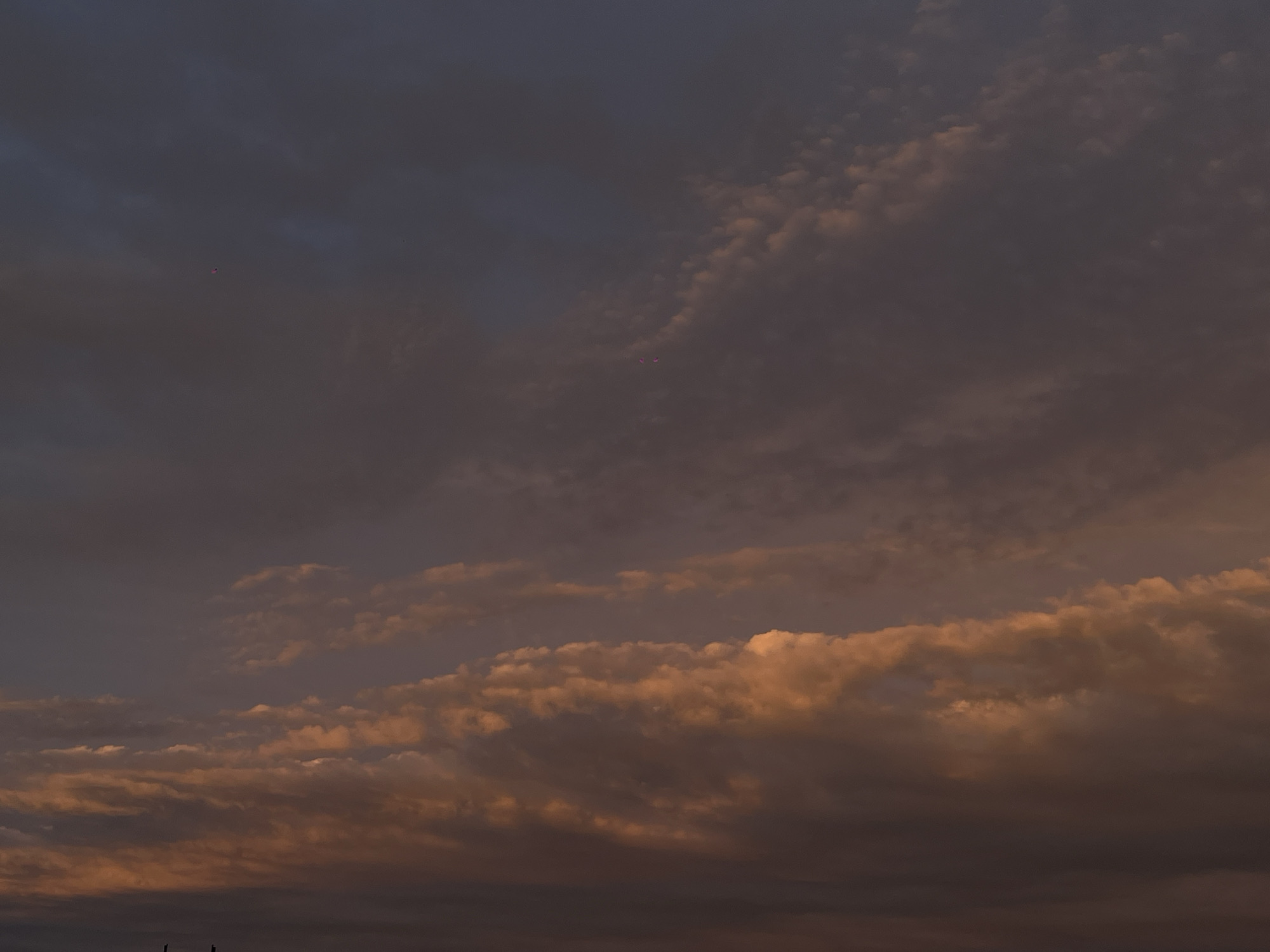 The image shows a dusky sky with dark, heavy clouds tinged with orange. The scene appears to be at dusk or dawn, as the light is dim. The location is possibly Novopokrovskaya, Russia, based on the prompt, but there are no identifying features in the image to confirm this. The horizon line is low, with a few dark shapes that may be trees or structures silhouetted against the sky.