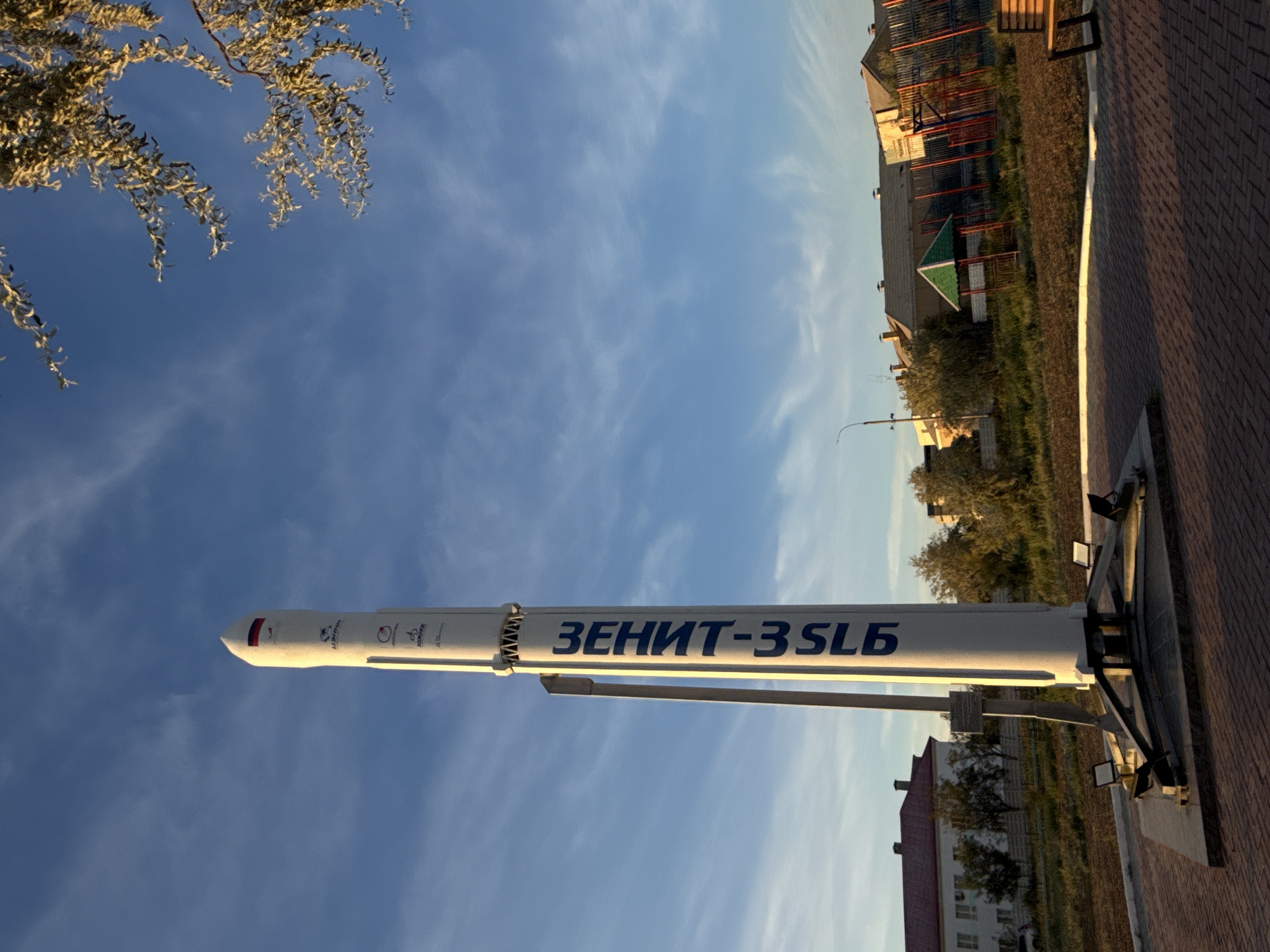 The image depicts a tall, white model rocket, identified by Cyrillic lettering as "ЗЕНИТ-3SLB," standing upright on a pedestal in what appears to be a public space within the city of Baikonur, Kazakhstan. The rocket model is adorned with several logos and a Russian flag decal near its nose cone. The surrounding environment features a paved area with brick-like tiles, a grassy patch with some shrubs and trees, and in the background, several buildings, a playground structure with a green roof, and a fence. The sky above is a clear blue with wispy clouds, illuminated by what seems to be late afternoon or early evening sunlight, casting long shadows. There are no people visible in the frame. The scene suggests a monument or display related to space exploration, common in Baikonur due to its historical association with the Baikonur Cosmodrome.