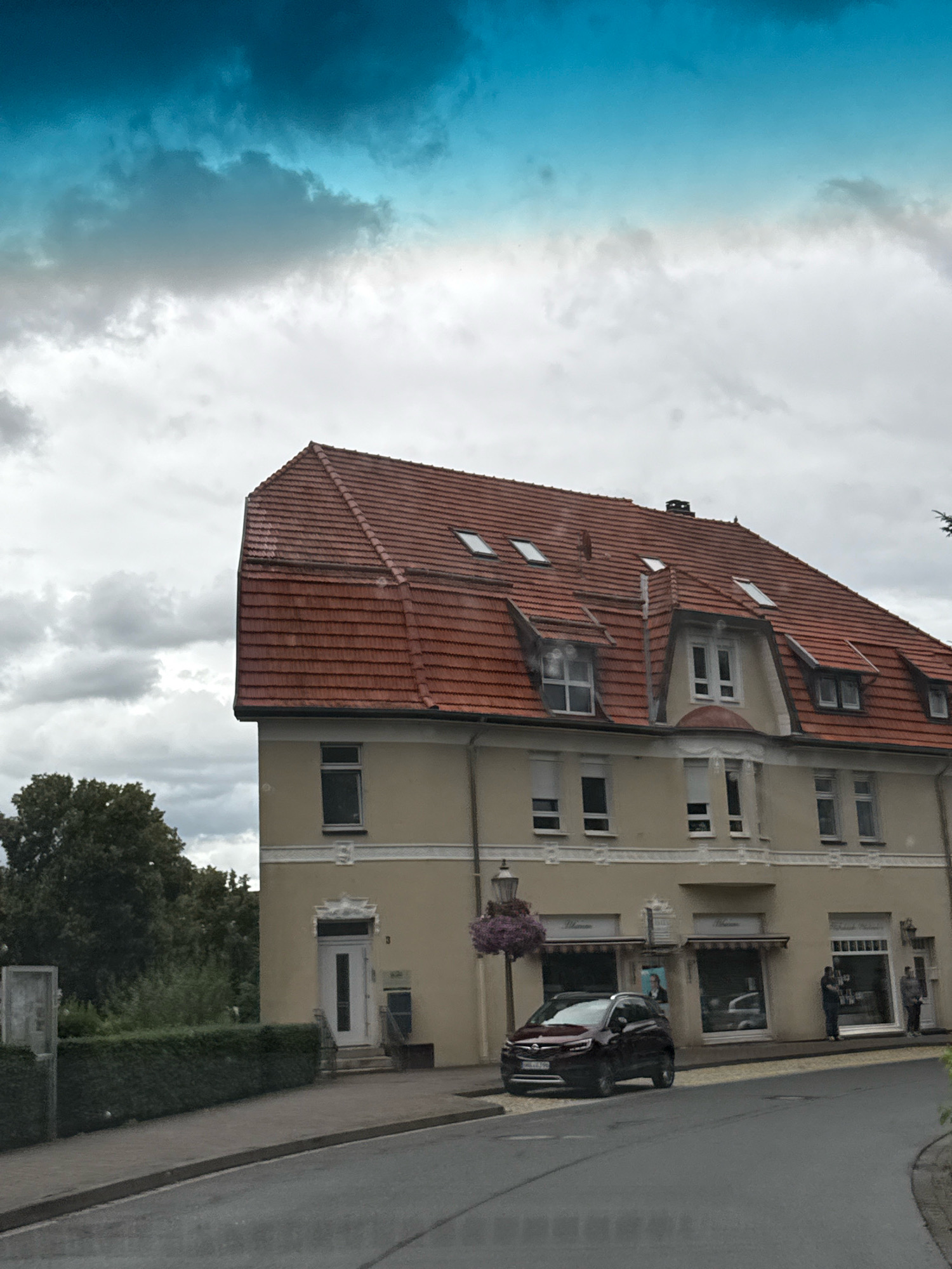 The image captures a view of a building in Bad Sachsa, Germany. The building has a light yellow facade and a red tiled roof with multiple dormer windows. A dark red car is parked on the street in front of the building. The street is paved and curves to the right. To the left of the building, there is a neatly trimmed hedge and a tree. In the background, the sky is overcast with dramatic clouds. The ground appears wet, suggesting recent rain. The building has several windows and storefronts on the ground floor, with awnings extended over them. Two individuals are visible standing near one of the storefronts.