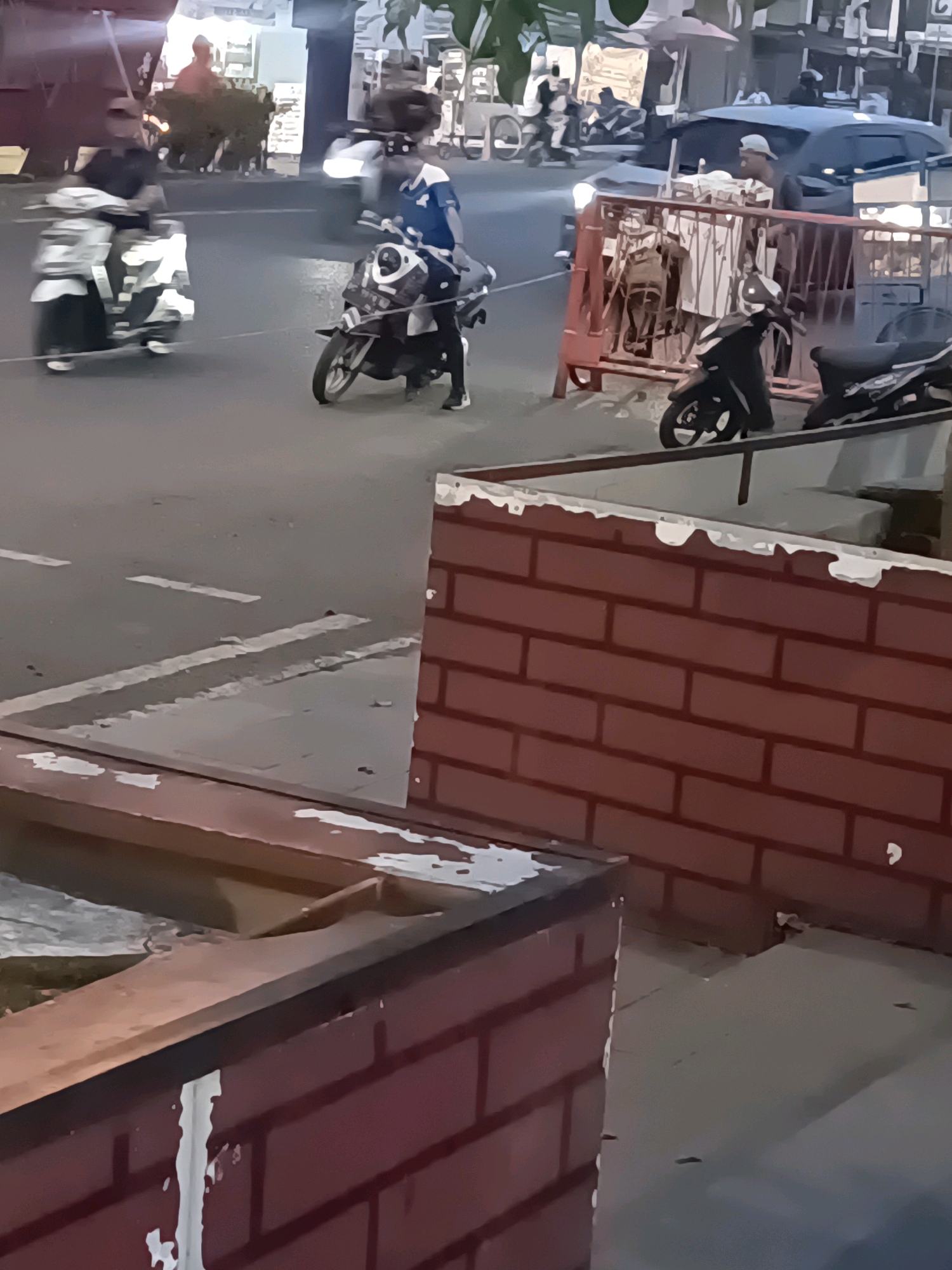 The image depicts a street scene in Lubuklinggau, Indonesia, likely during dusk or early evening given the lighting conditions. In the foreground, a textured reddish-brown brick wall and a concrete ledge create a layered foreground. The middle ground features a road with visible white lane markings and a crowd of people on foot and riding motorcycles.
Several motorcycles are in motion: one is prominently in the center right, a rider in a blue shirt and black pants is on it, wearing a helmet. To the left, another motorcycle with a rider wearing dark clothing is moving, with a third scooter further left, also with a rider. A black car is visible behind a red metal barricade in the middle ground, and a person wearing a light-colored cap and dark shirt is standing near the barricade. Several stalls or vendor carts are visible further in the background. The overall impression is a bustling urban environment with typical street traffic and activity.