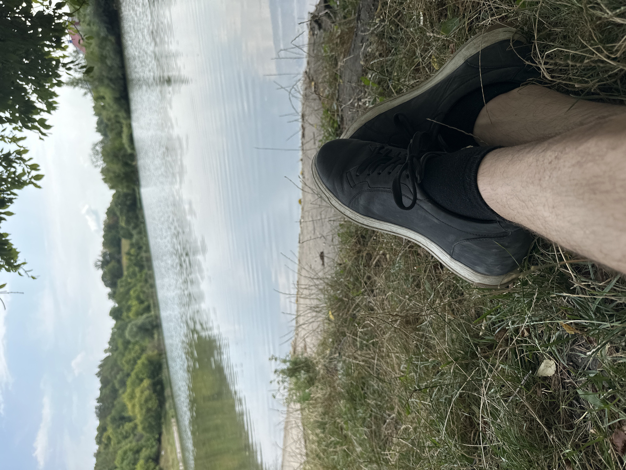 The image shows a first-person perspective of a person sitting on the grassy bank of a body of water, with their legs crossed and feet in black sneakers. The water is calm with ripples, reflecting the sky and the tree-lined shore. The environment appears to be a natural setting, possibly a lake or a wide river, with lush green trees and vegetation on the opposite bank. A concrete structure runs along the edge of the water. The sky is partly cloudy, indicating daylight. The location is identified as Makiv, Ukraine, suggesting a rural or semi-rural landscape. There is no visible interaction between people or specific actions being performed beyond the act of sitting and resting. The overall impression is one of peacefulness and relaxation in nature.