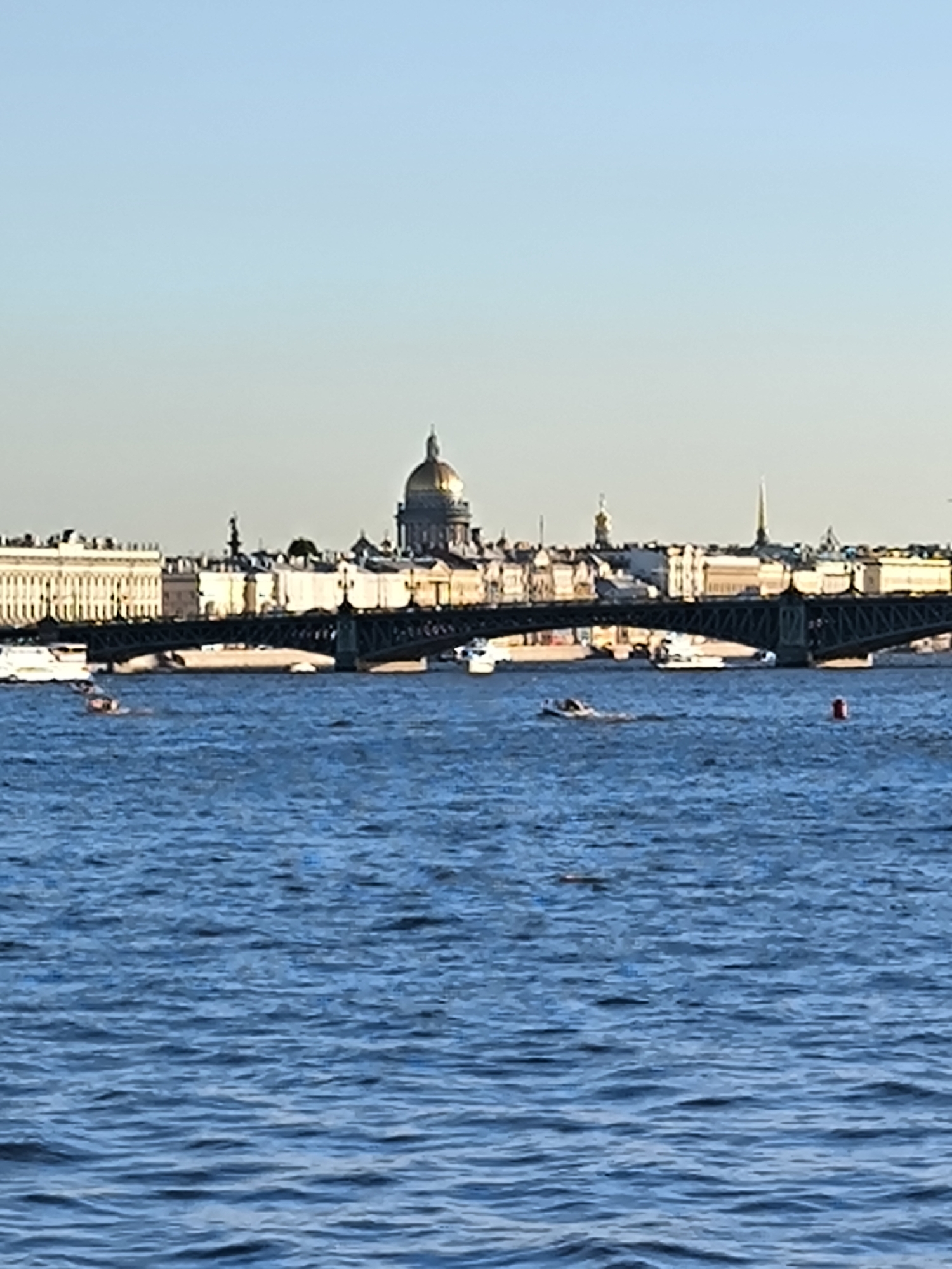 A wide shot captures a cityscape on a clear, bright day with a deep blue sky. In the foreground, a body of water, likely a river or canal, is filled with ripples and waves. Several boats are visible on the water, including a white motorboat to the left, and a smaller, darker boat with passengers in the center. A red buoy is also present in the water on the right.
Dominating the midground is a long, dark bridge with multiple arches, spanning across the water. Beyond the bridge, the St. Petersburg skyline is visible. Prominent structures include grand, light-colored buildings with classical architecture. The most striking feature is a large, golden-domed cathedral, likely St. Isaac's Cathedral, which stands tall against the sky. Several other spires and towers dot the skyline, indicative of a historic European city. The buildings appear to be illuminated by the sun, casting subtle shadows. The overall impression is one of a beautiful, sunlit day in a historic Russian city.
