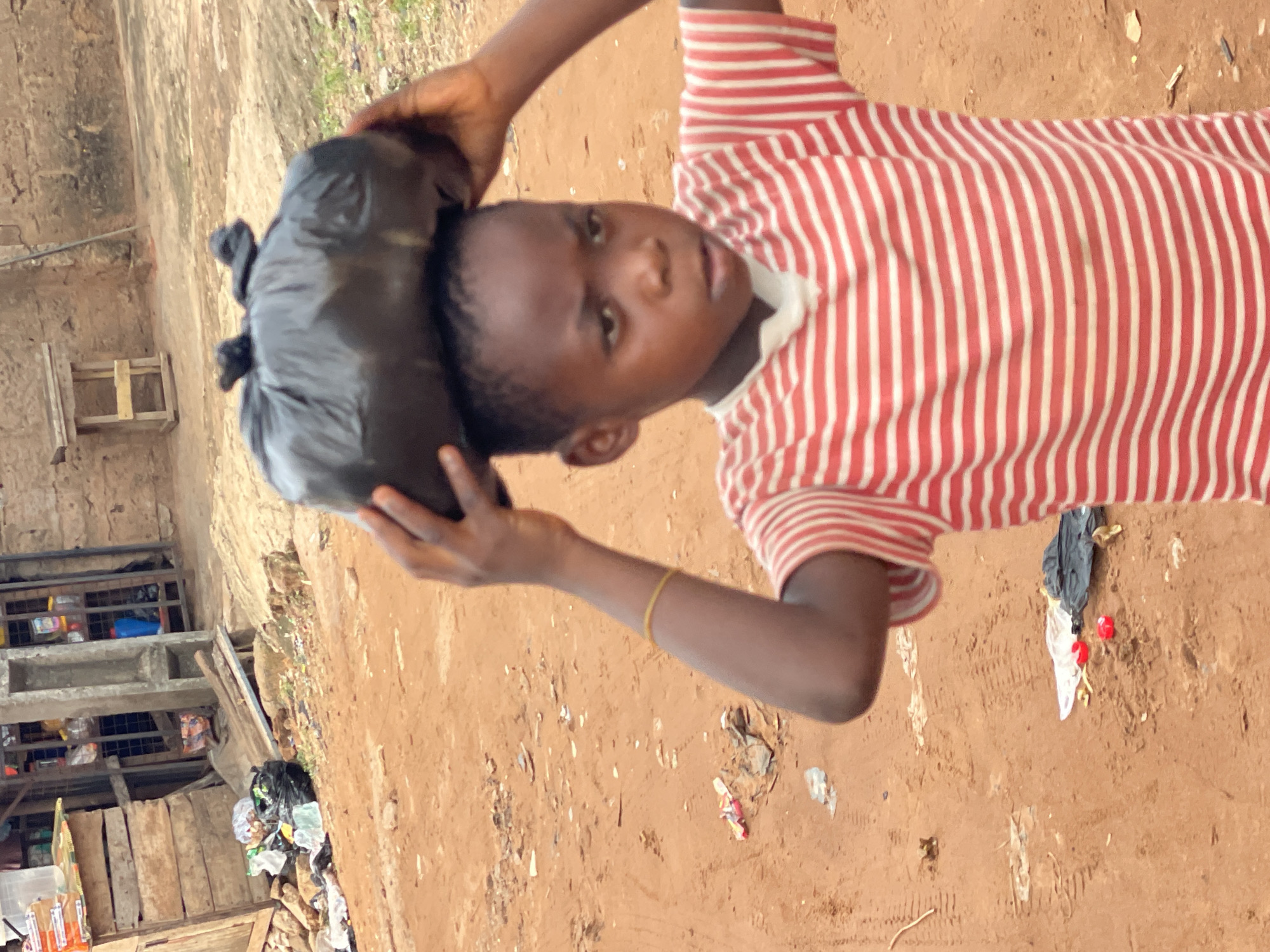 A child wearing a red and white striped shirt carries a black plastic bag on their head. The location appears to be Onitsha, Nigeria, due to the presence of a dirt ground, buildings with stalls and general waste in the environment. The child is facing the camera. The background is blurry, with building details including wooden support structures, and garbage visible. The ground is brown.