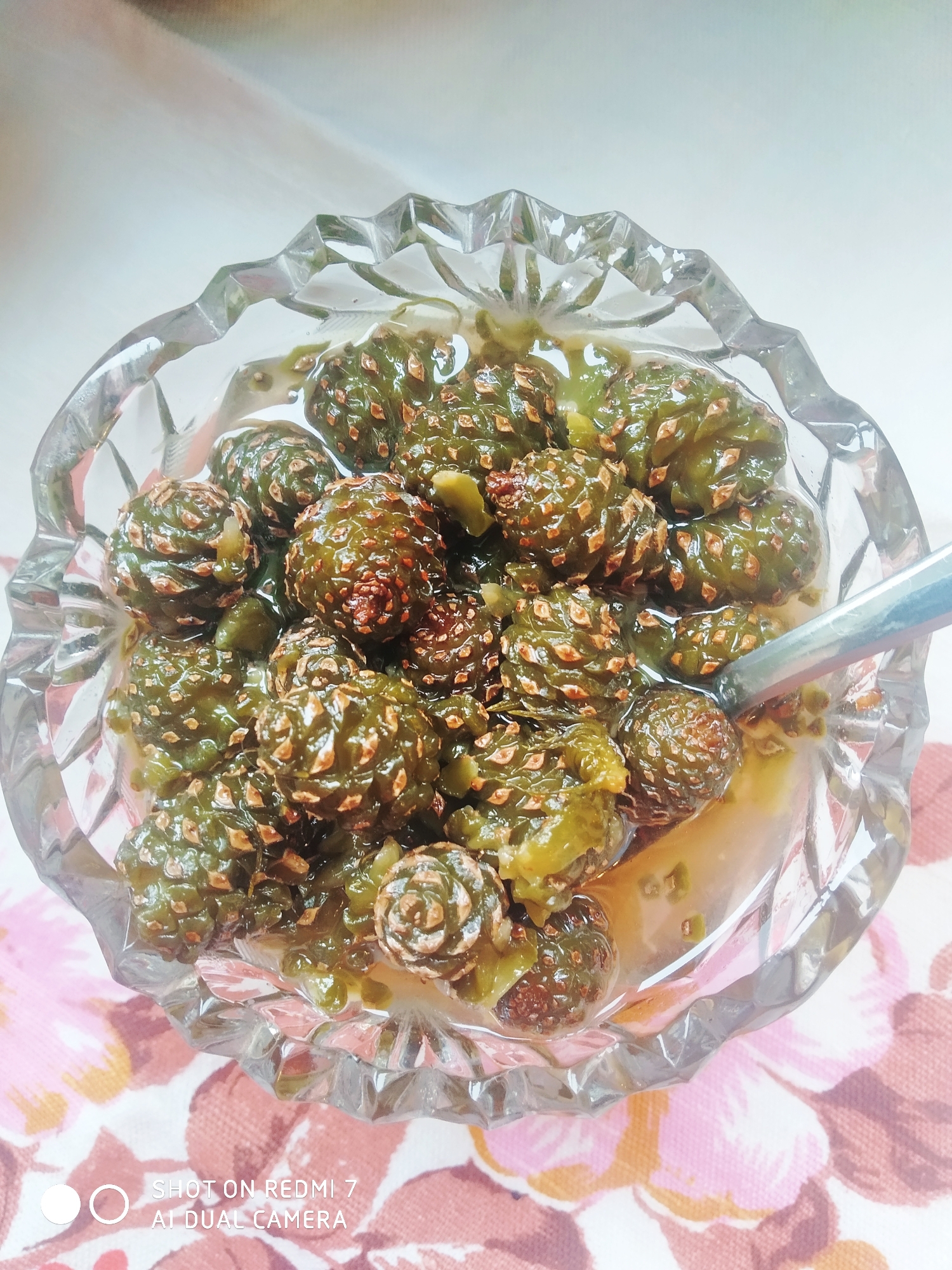 A close-up, high-angle shot shows a cut-glass bowl filled with what appears to be pine cone jam on a floral-patterned tablecloth. A spoon rests in the jam. The jam consists of numerous small, green and brown pine cones preserved in a translucent, amber-colored syrup. The bowl has a scalloped, decorative edge. "SHOT ON REDMI 7 AI DUAL CAMERA" is visible in the lower left corner of the image, suggesting the photograph was taken with a Redmi 7 smartphone. There is no visible evidence that the image was taken in Chaikovsky, Russia.
