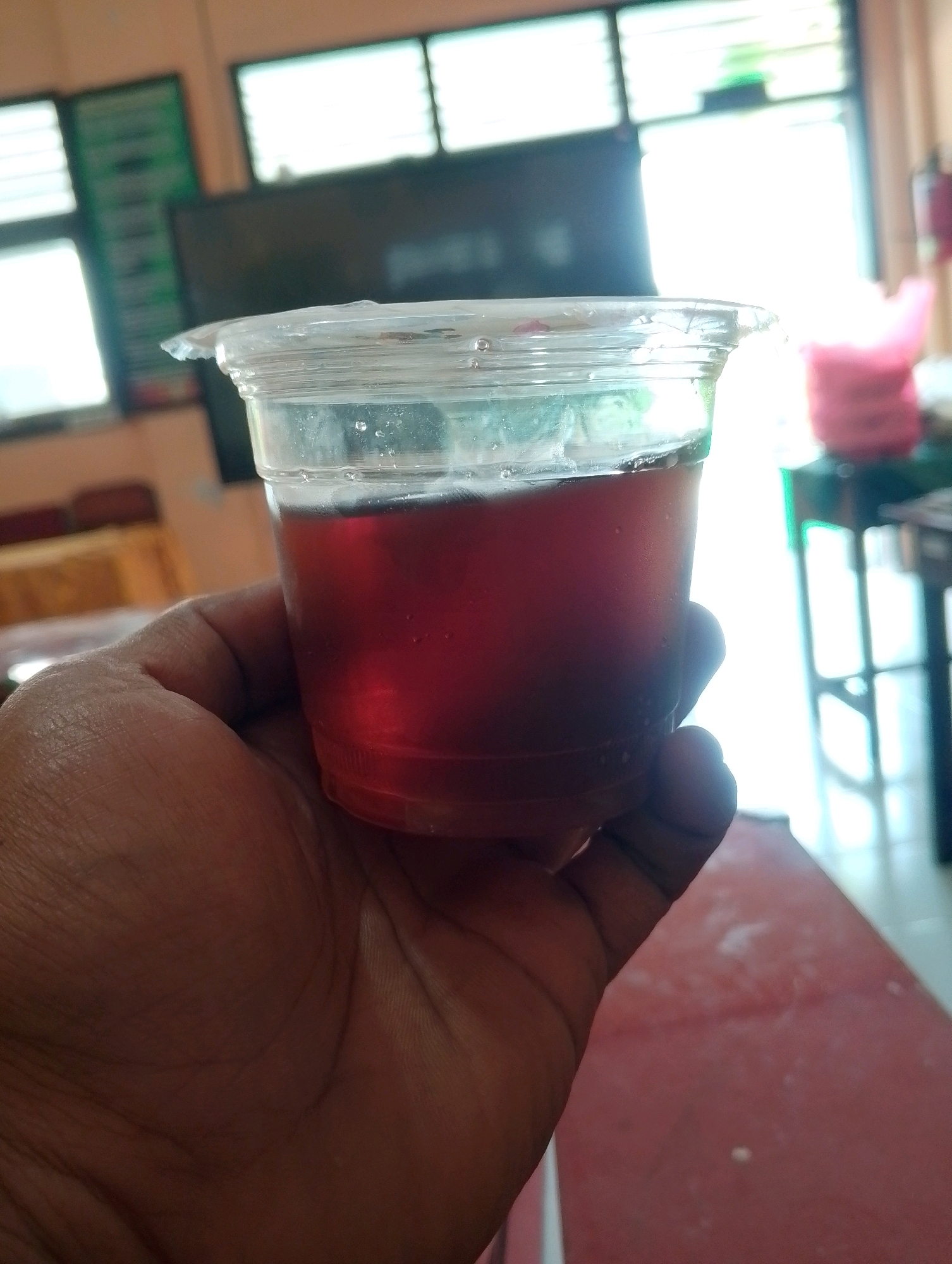 A close-up shot shows a hand holding a clear plastic cup filled with a dark red liquid, possibly iced tea or juice. The cup has condensation on its exterior and a slightly damaged lid. The background is blurred, but suggests an indoor setting with windows showing bright daylight. A red table surface is visible in the foreground. The scene appears to be in Papungan, Indonesia.