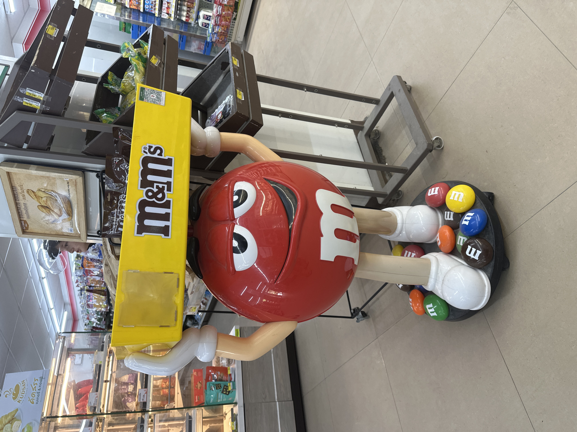 The image captures a large M&M's character display positioned in what appears to be a convenience store, likely a 7-Eleven, in Koh Phangan, Thailand. The character, a red M&M, is holding a yellow M&M's branded display box above its head. Its arms are white and bulbous, and its feet are also white, standing on a circular base adorned with various colored M&M's candies, each featuring a white "m" logo. The display is situated on a light-colored tiled floor, common in commercial establishments.
Behind the M&M's character, shelves are visible, stocked with various products. To the left, refrigerated display cases contain packaged food items. To the right, open shelves hold more merchandise, including what looks like bags of snacks and bottles. The overall lighting suggests an indoor environment, typical of a retail space. A person wearing a clear plastic face shield is partially visible in the background, standing near the shelves, suggesting they are either a customer or an employee. The presence of M&M's branding and the store's layout strongly suggest a retail setting within Koh Phangan.