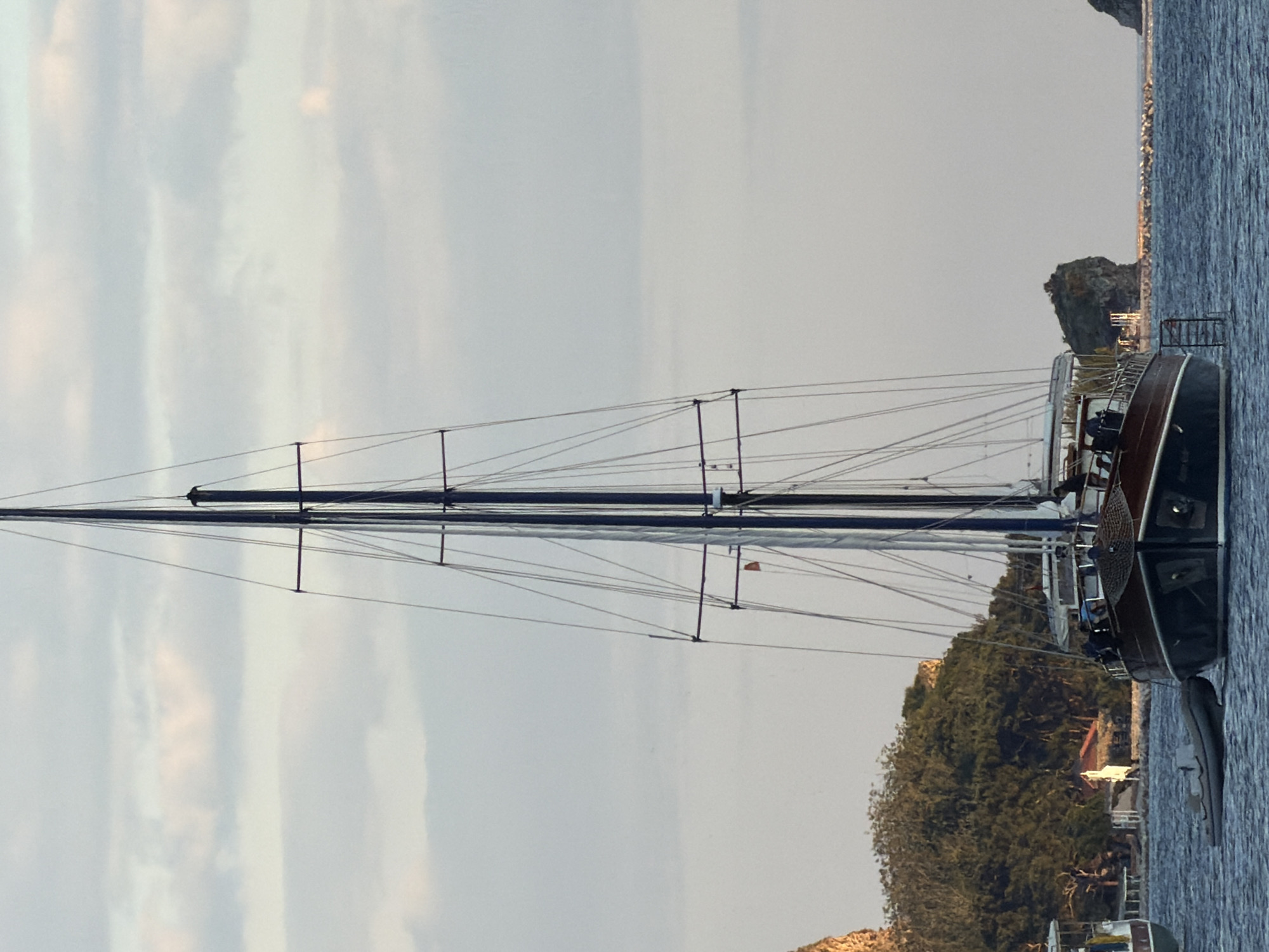 The image shows a sailing yacht moored in a body of water, possibly a bay or harbor. The yacht is a large sailing vessel with a tall mast and numerous rigging lines. The hull of the yacht appears to be a dark color. There are other boats nearby, and land is visible in the background. The land is covered with trees and vegetation. There is a rocky outcrop also visible. The sky is overcast. The location is Unknown, Unknown.