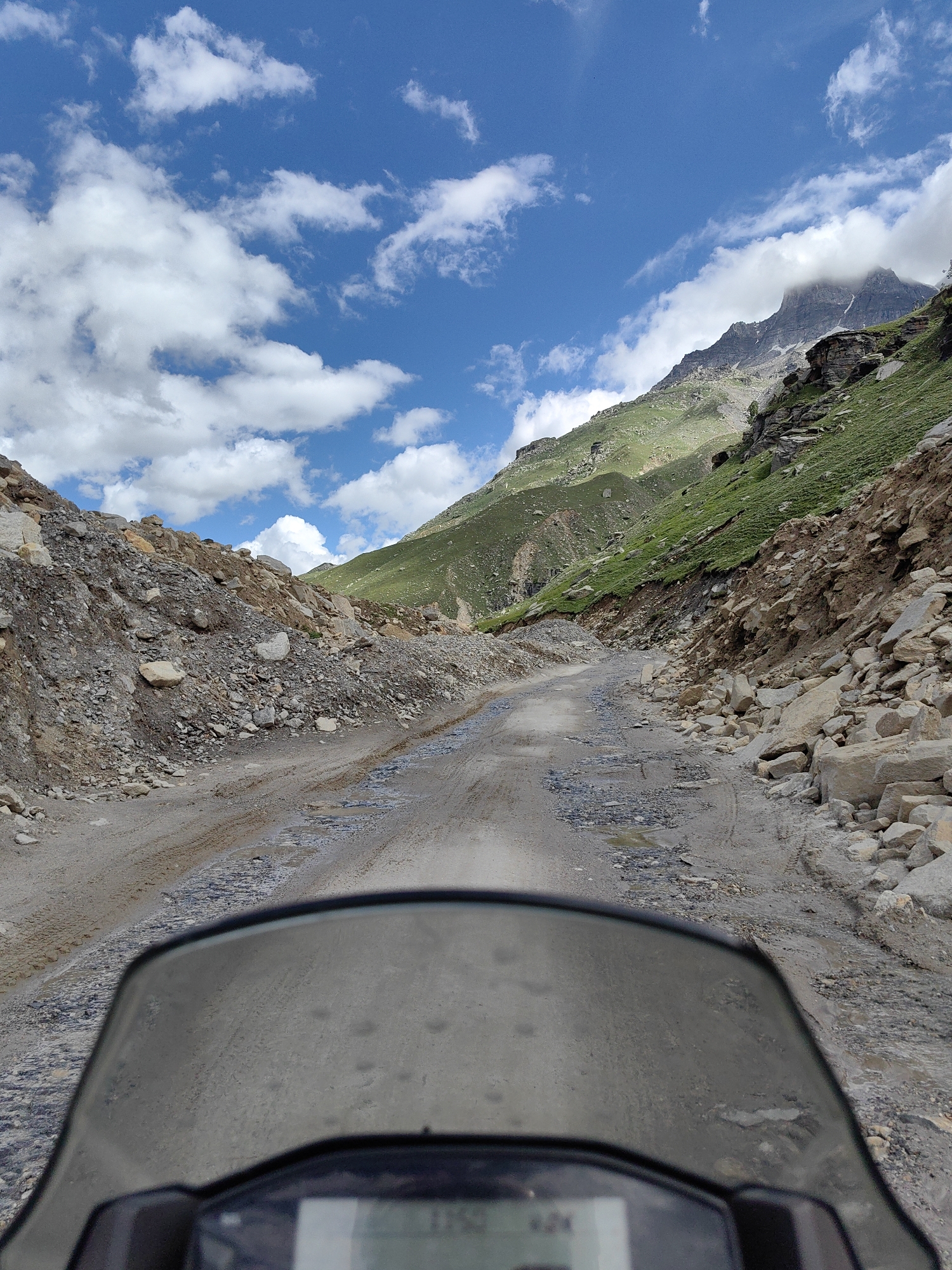 The image shows a point-of-view shot from a motorcycle on a dirt road in the mountains. The road is narrow and appears to be unpaved, with loose gravel and some puddles of water. The surrounding landscape is mountainous, with steep, grassy slopes on the right and rocky, barren slopes on the left. The sky is blue with scattered white clouds. The motorcycle's windshield and dashboard are visible in the foreground. The location is identified as Bashisht, India.