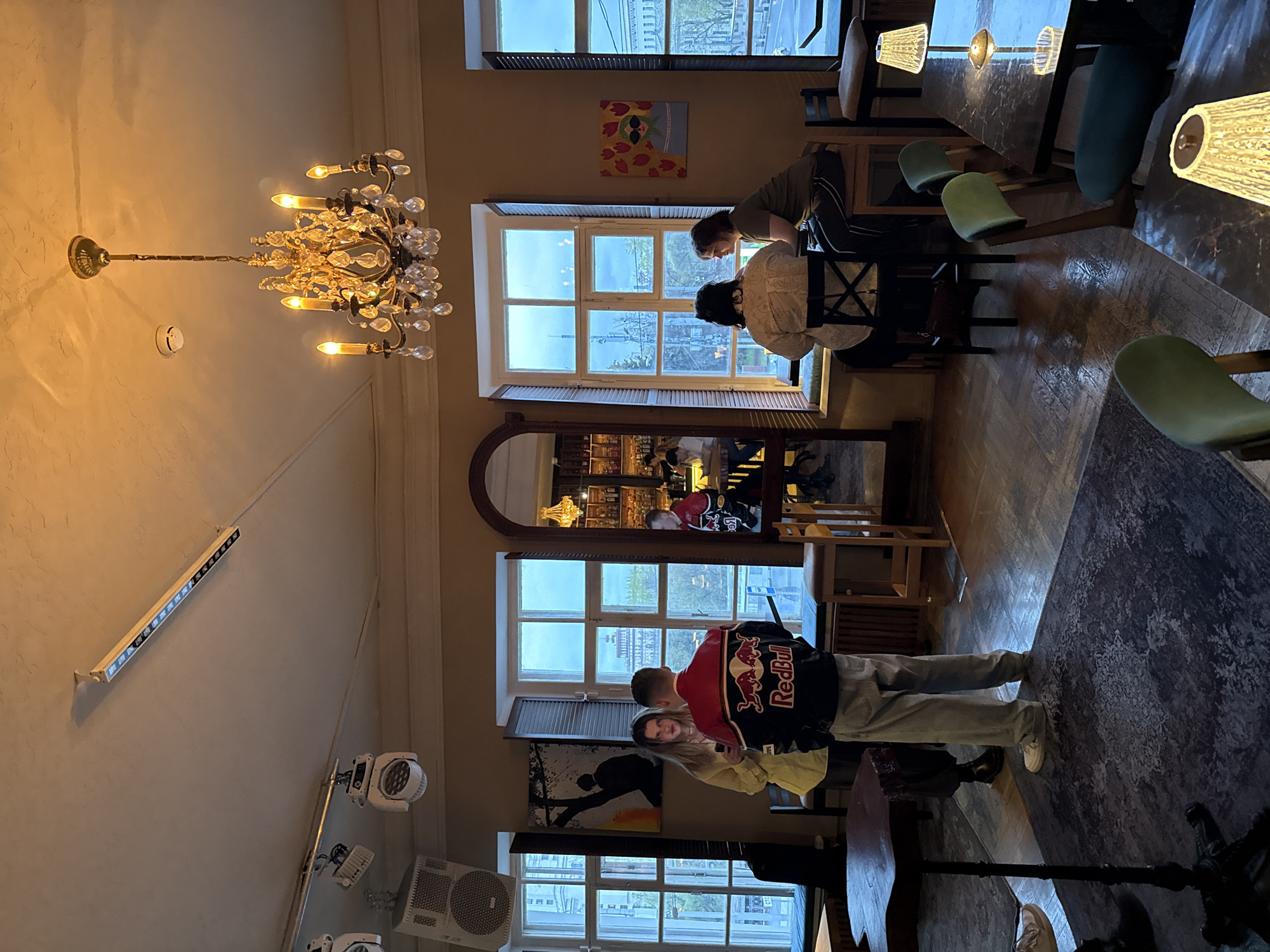 The photo shows the interior of a restaurant in Moscow, Russia. The restaurant has tables and chairs, and people are seated at some of the tables. There is a chandelier hanging from the ceiling. A couple stands near a window, and one of them is wearing a Red Bull jacket. There are paintings on the wall. The restaurant has large windows that provide natural light.