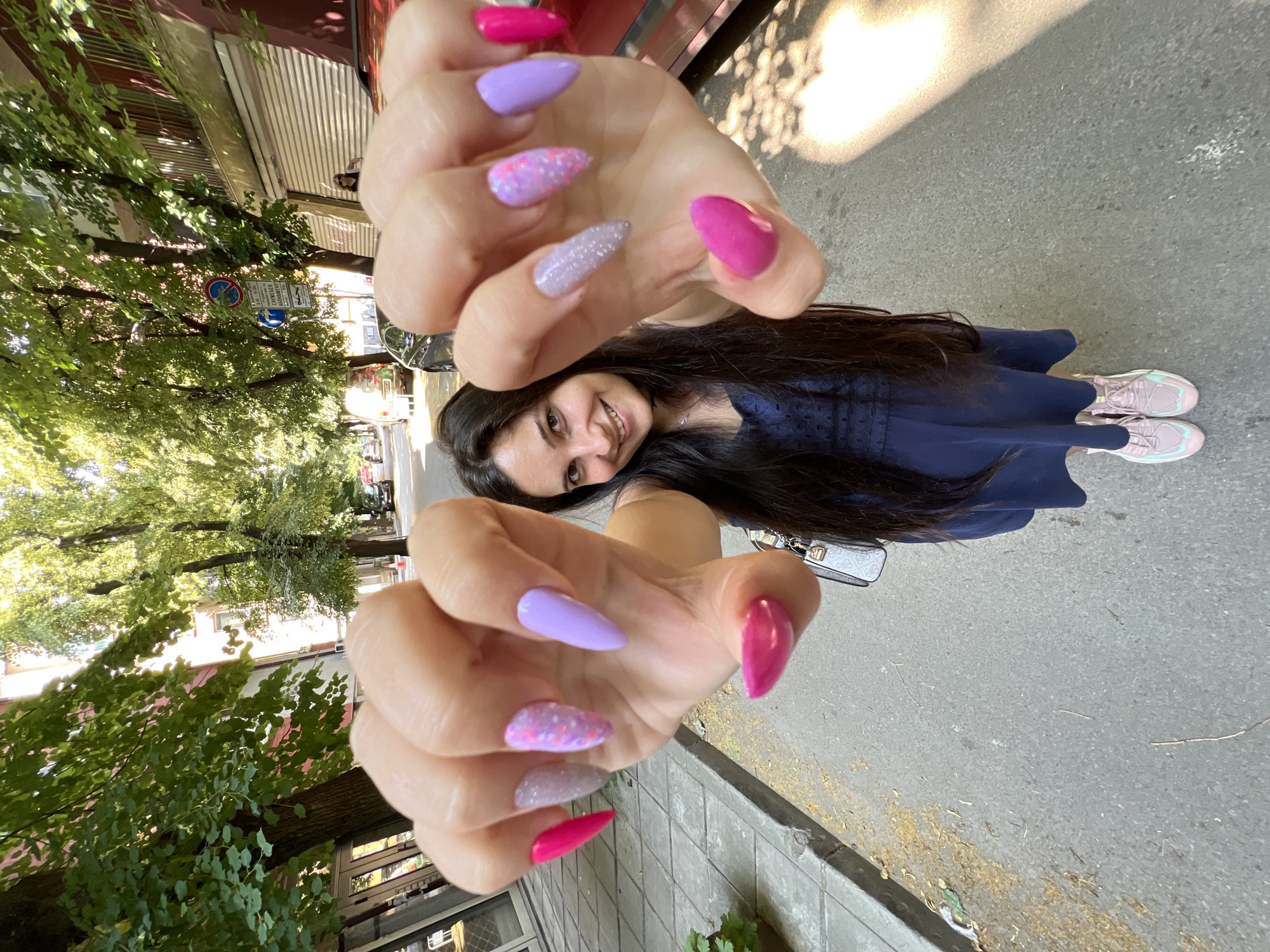 This is a photograph of a young woman with long, dark hair, wearing a navy blue dress and pink sneakers. She is smiling at the camera and holding her hands up, displaying her fingernails which are painted in various shades of pink and purple, some with glitter. The woman is standing on a paved sidewalk next to a tree-lined street. In the background, there are buildings and parked cars. The image appears to have been taken during the daytime, as evidenced by the sunlight casting shadows on the ground. The presence of trees and buildings suggests an urban or suburban environment. There are no visible signs of specific weather conditions, but the lighting indicates it is likely a sunny day. The overall mood of the image is cheerful and relaxed.