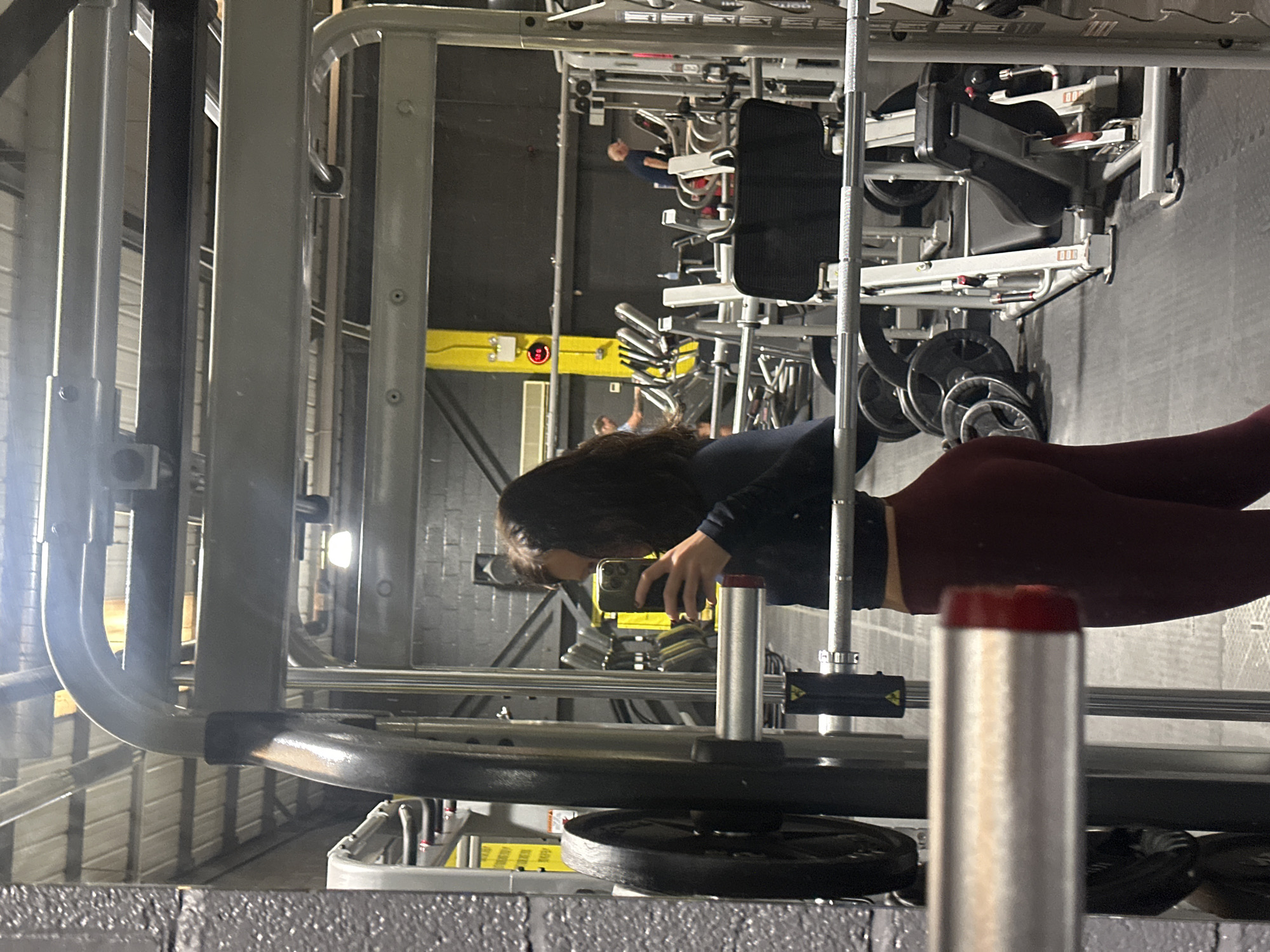 The image is taken inside a gym, probably in Tonbridge and Malling, United Kingdom, with gym equipment prominently visible. A person with dark hair is holding a phone up to take a picture. The person is wearing dark clothes and burgundy colored leggings. The focus of the photograph seems to be on the gym equipment, including a weight rack loaded with weight plates and a machine with adjustable seats and levers in the background. Other gym equipment is visible further back in the image. The environment is brightly lit and appears well-maintained. Another person can be faintly seen using a machine in the distance.
