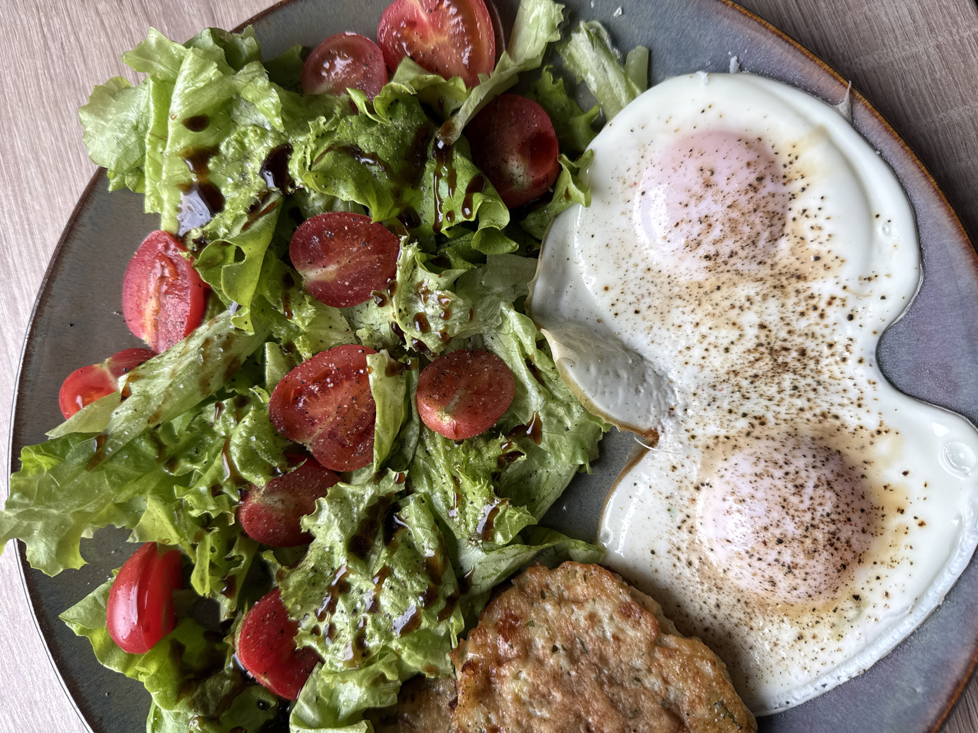 A top-down view shows a grey plate on a wooden surface, likely a table. The plate holds a meal consisting of a salad, two fried eggs, and what appears to be a sausage patty. The salad is made of lettuce and sliced cherry tomatoes, drizzled with a dark balsamic glaze. The eggs are fried sunny-side up, with pepper sprinkled on top. The sausage patty is browned and slightly textured. The image does not include any information about the location in Krasnoyarsk, Russia.