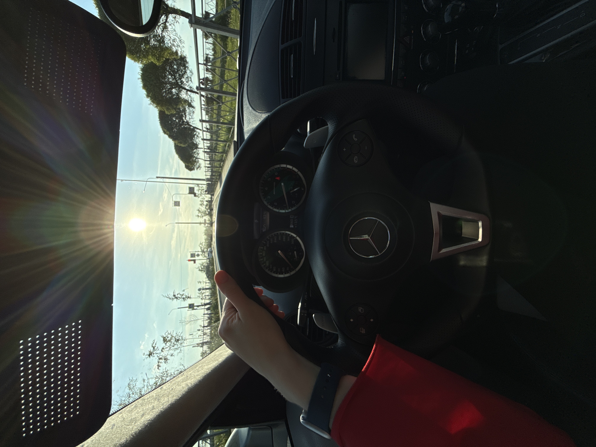 The image captures a first-person perspective from the driver's seat of a car on a bright, sunny day, likely in Saint Petersburg, Russia. A person's right hand, adorned with a dark blue Apple Watch, grips the steering wheel. A red sleeve is visible on the arm.
The car's interior features a black leather steering wheel with the prominent Mercedes-Benz logo at its center. Behind the steering wheel, the instrument cluster is partially visible, showing circular gauges with green backlighting, indicating information such as speed and engine RPM. Digital readouts within the cluster display "P" (for Park), "93°F" (likely ambient temperature), and "105°C" (possibly engine temperature). The dashboard, primarily dark, includes air vents and a portion of the central console.
Through the windshield, a clear blue sky dominates the exterior view. The sun is low in the sky, creating a powerful glare and lens flare on the left side of the frame, indicating either early morning or late afternoon. The visible landscape includes a tree-lined road or parkway with lush green trees and a metallic fence or guardrail. Street lights and a traffic light structure are visible in the distance. A distinct perforated pattern is present on the left side of the windshield, possibly a built-in sunshade or tint feature. The scene conveys a calm moment during a drive or while stopped, under clear weather conditions.